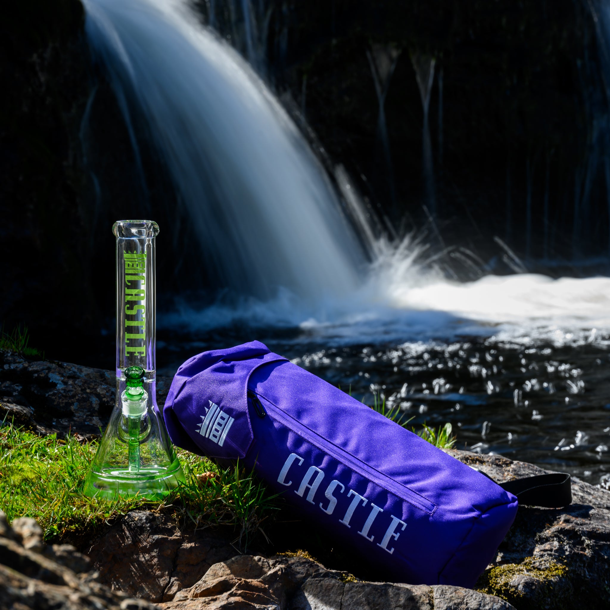 A sleek glass bong from Castle Glassworks, featuring bold green branding, stands on a rocky surface near a waterfall. Next to it lies a vibrant purple Double Layer bag with the "CASTLE" logo, adding a stylish and functional touch. The natural backdrop of flowing water and lush greenery enhances the aesthetic, creating a contrast between rugged outdoor elements and the refined craftsmanship of the glass piece. The scene exudes adventure and quality, highlighting the bong's portability and premium design.