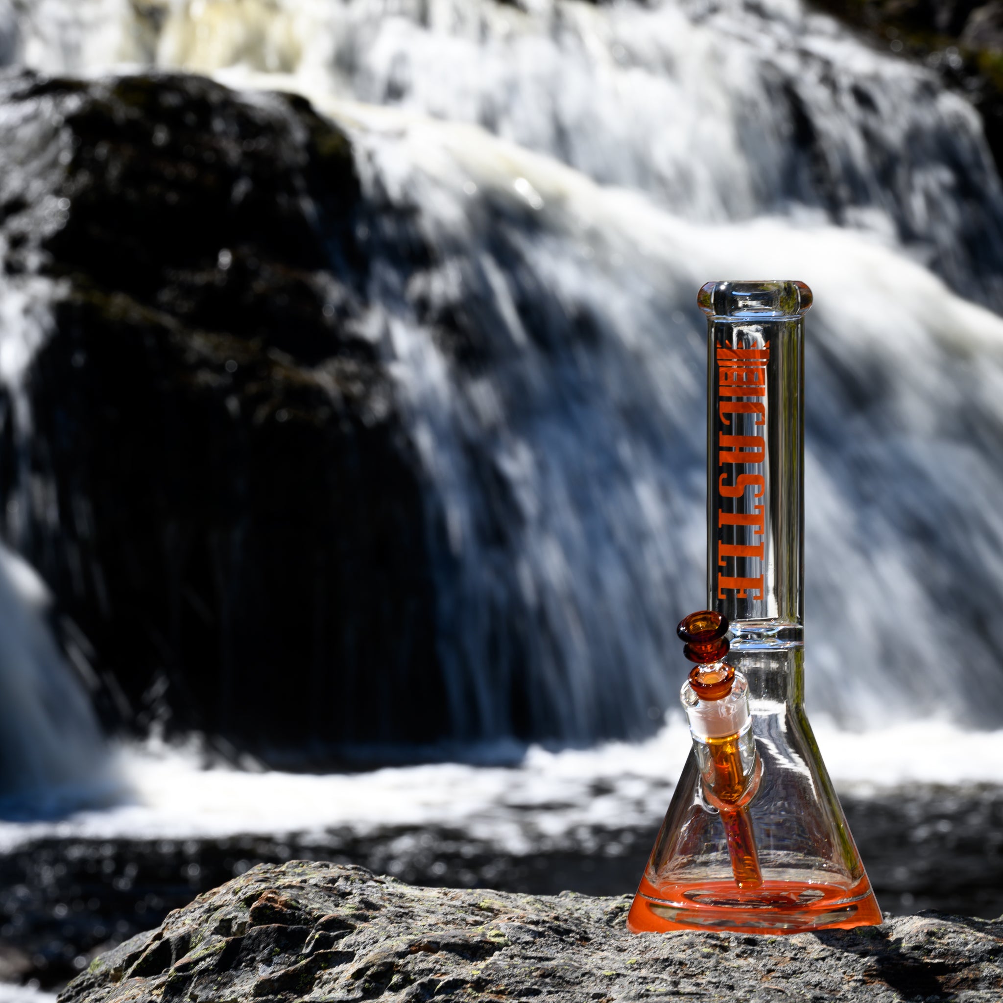 A striking glass bong from Castle Glassworks with bold amber accents is placed on a rugged rock surface in front of a rushing waterfall. The transparent beaker-style piece features vibrant "CASTLE" branding on the neck, a matching orange downstem, and a glass bowl with amber hues. The powerful flow of water in the background enhances the dynamic and adventurous feel of the image, emphasizing the craftsmanship and durability of the bong in an outdoor setting.