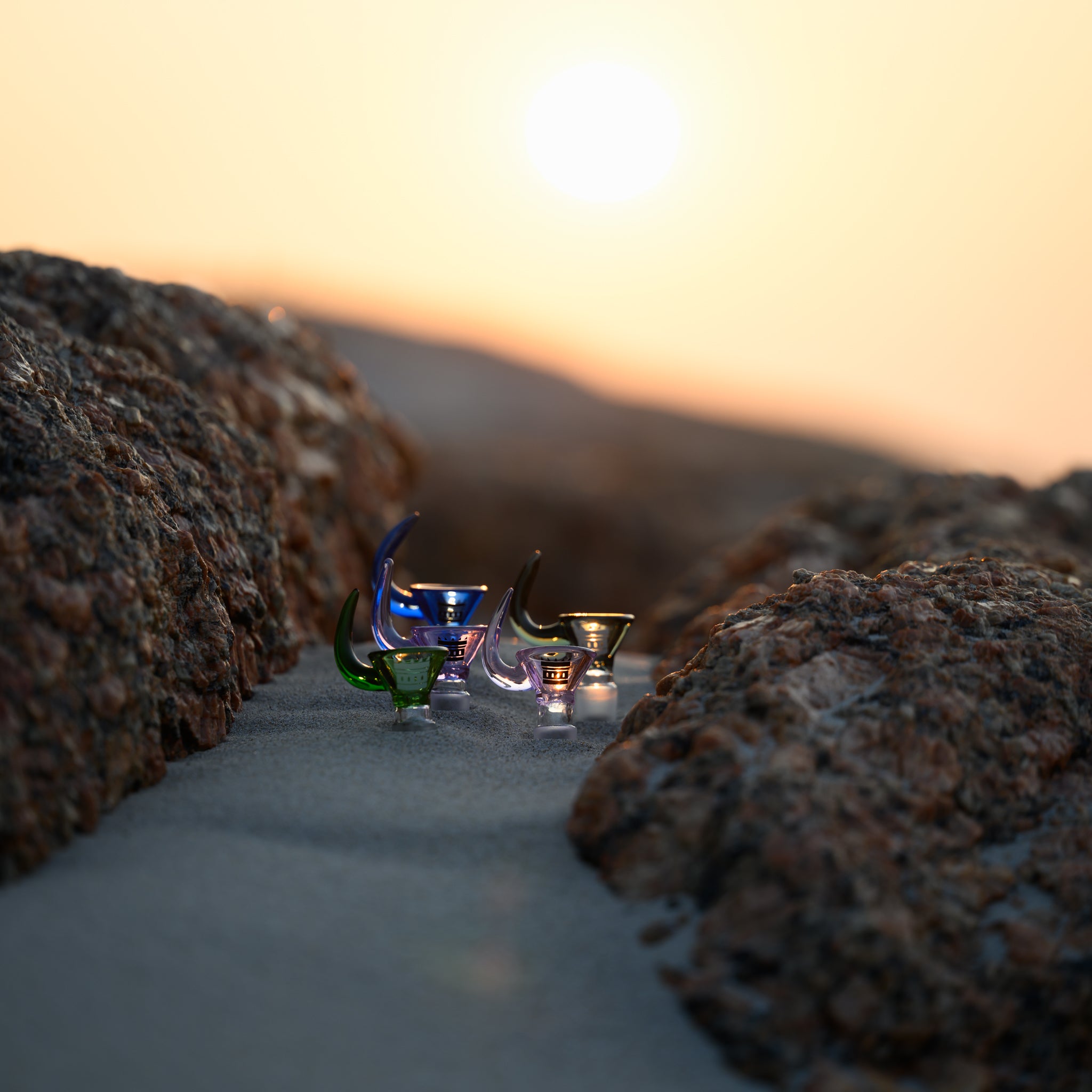 A set of glass horn bowls in various colors, including green, blue, and purple, displayed on a sandy surface between rugged rocks at sunset. The soft golden light highlights the intricate design and smooth curves of the glass.