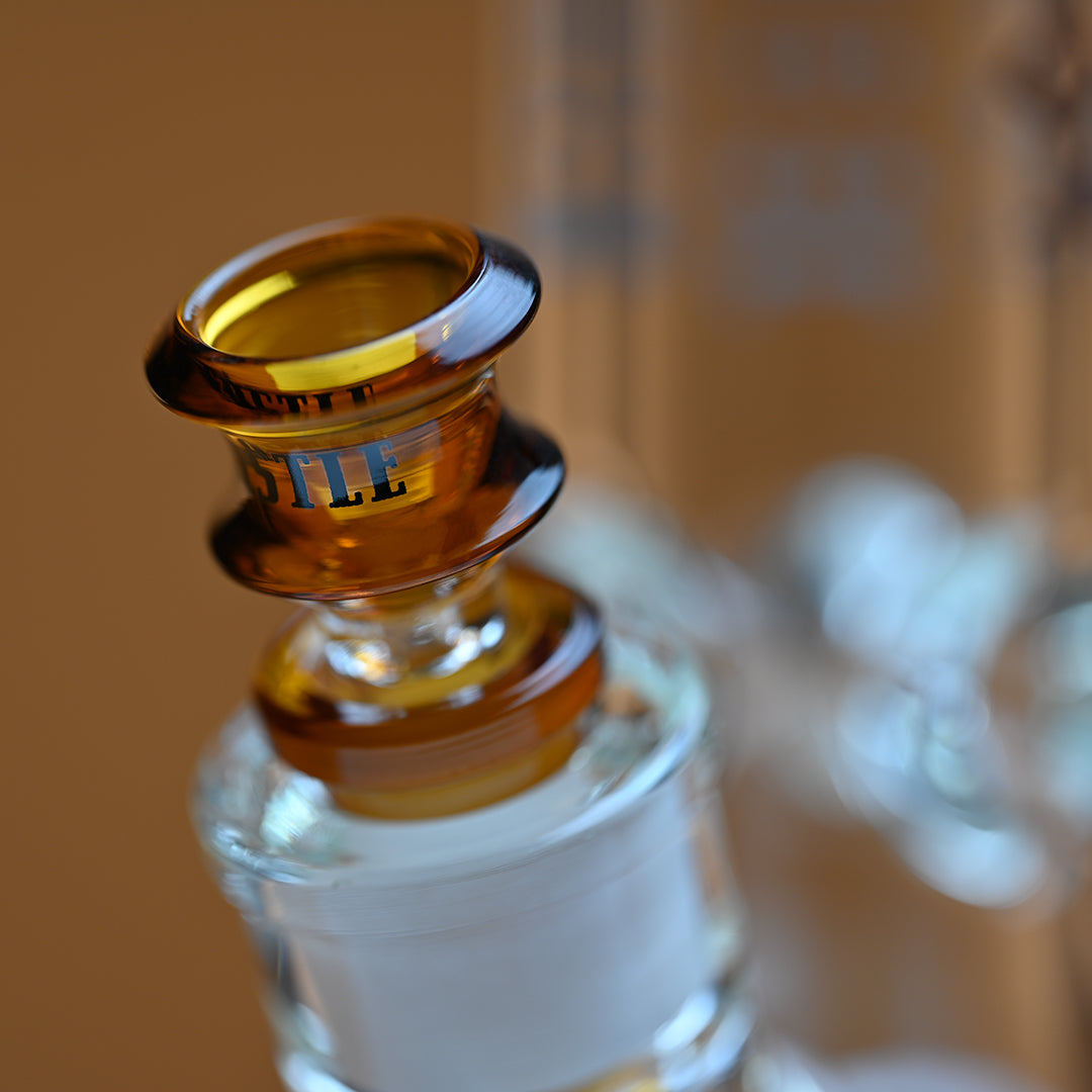 A close-up image of an amber colored glass bowl attached to a bong, with the brand name Castle in black letters on the side of the bowl. The rich amber color of the glass contrasts beautifully with the transparent glass of the bong, and the background is softly blurred in a warm amber hue. The focus on the bowl highlights the craftsmanship and detail, giving the image a refined and elegant appearance.