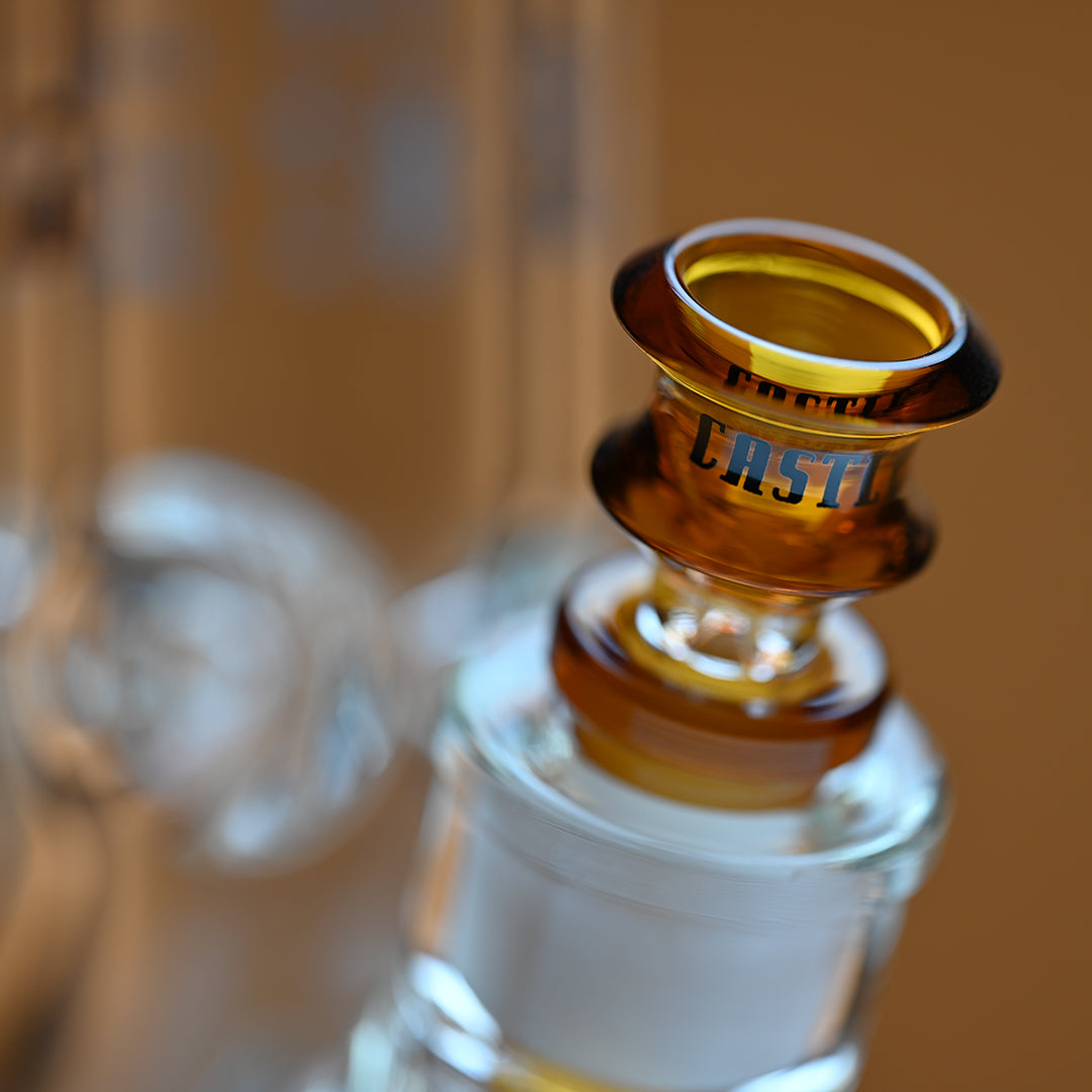 A close-up image of an amber-colored glass bowl attached to a bong, with the brand name Castle in black letters on the side of the bowl. The image focuses on the intricate details of the bowl, with the amber glass reflecting the light beautifully. The background is softly blurred in a warm amber tone, complementing the color of the bowl and enhancing the overall warmth and elegance of the scene.