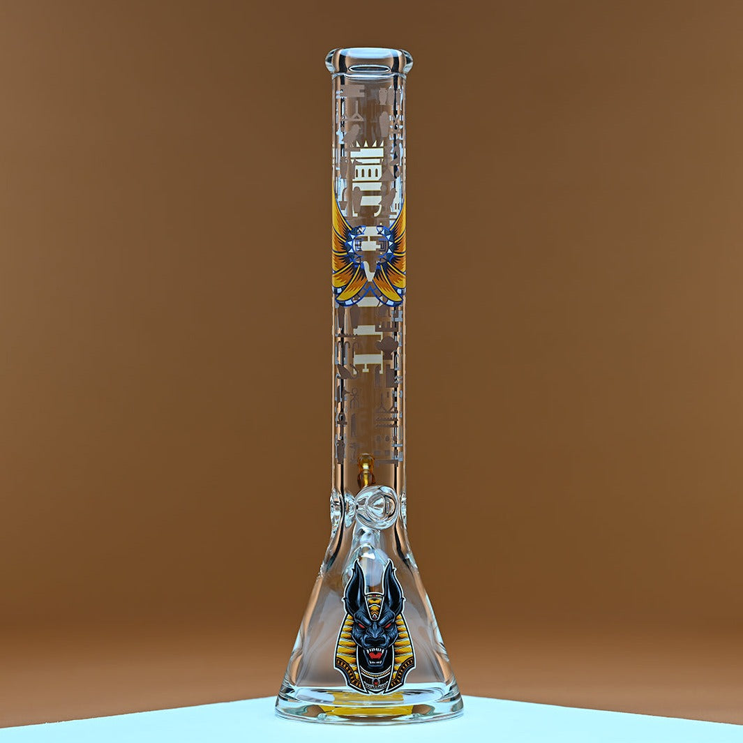 A glass bong with yellow accents and the brand name Castle in bold yellow letters on the neck, featuring an intricate Anubis design on the base. The bong is placed on a surface with a clean, minimalist background in warm amber tones, creating a striking contrast that highlights the bong's sleek design and detailed artwork. The bong has an amber colored bowl and downstem, adding to its overall aesthetic appeal.