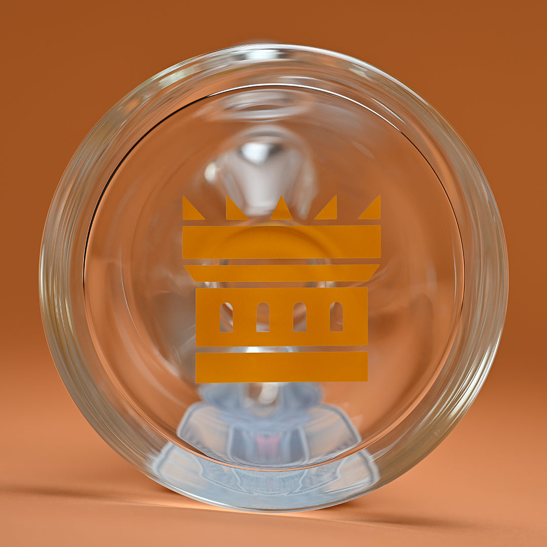 A close-up view of the bottom of a glass bong, showing the base with a bold, stylized castle logo in amber. The glass is transparent, allowing light to reflect off its surface, creating a sleek and polished appearance. The warm amber background complements the amber logo, enhancing the overall visual appeal and emphasizing the branding.