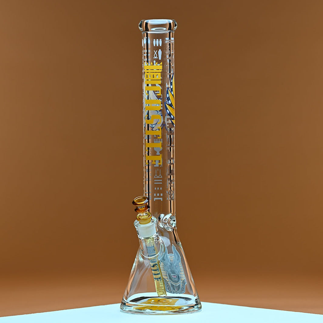 A glass bong with yellow accents and the brand name Castle in bold yellow letters, featuring Egyptian hieroglyphics and an intricate Anubis illustration on the base. The bong is positioned against a warm amber background, which contrasts with the clean, transparent glass and highlights the detailed design elements.
