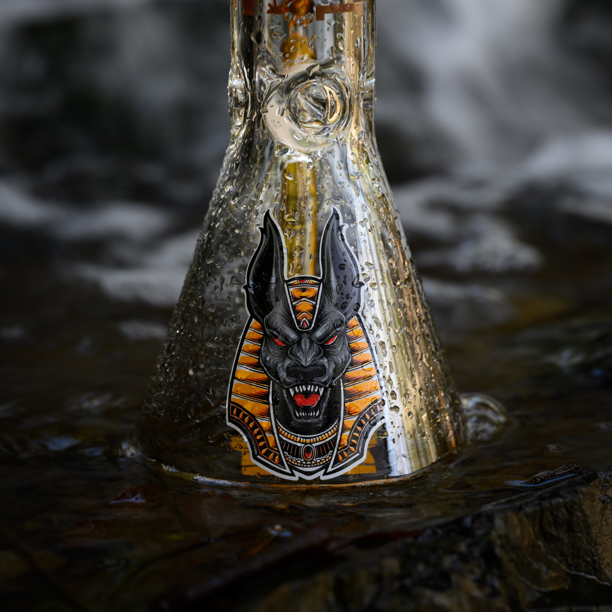 A clear glass bong with water droplets on its surface, featuring a detailed and fierce illustration of Anubis on the base. The bong is positioned in a natural setting with a flowing waterfall in the blurred background, enhancing the scene's dynamic and refreshing ambiance. The Anubis design is prominent, with intricate details and vibrant colors, standing out against the clear glass and natural surroundings.