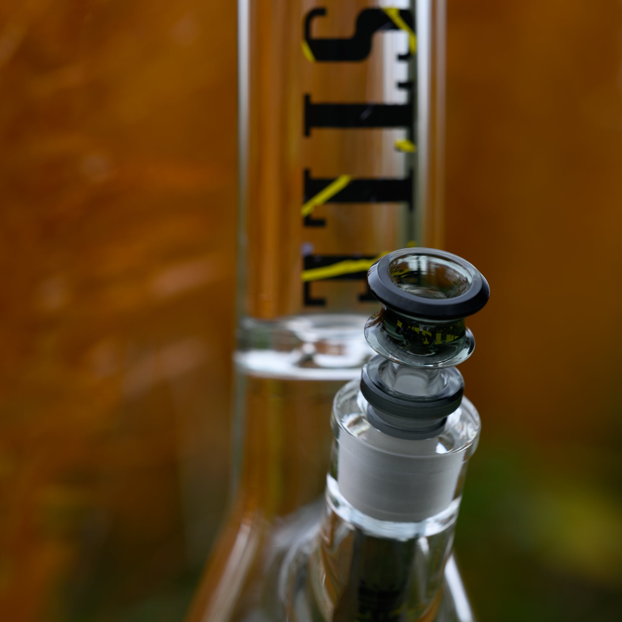A close-up of a high-quality glass bong from Castle Glassworks, focusing on the intricate details of the bowl and downstem. The bowl features a sleek black-rimmed design with a multi-ring structure, packed with finely ground material. The clear glass body showcases the "CASTLE" branding in bold black and yellow lettering. The blurred background of golden-hued grass creates a warm, natural contrast, emphasizing the precision craftsmanship and premium aesthetics of the piece.