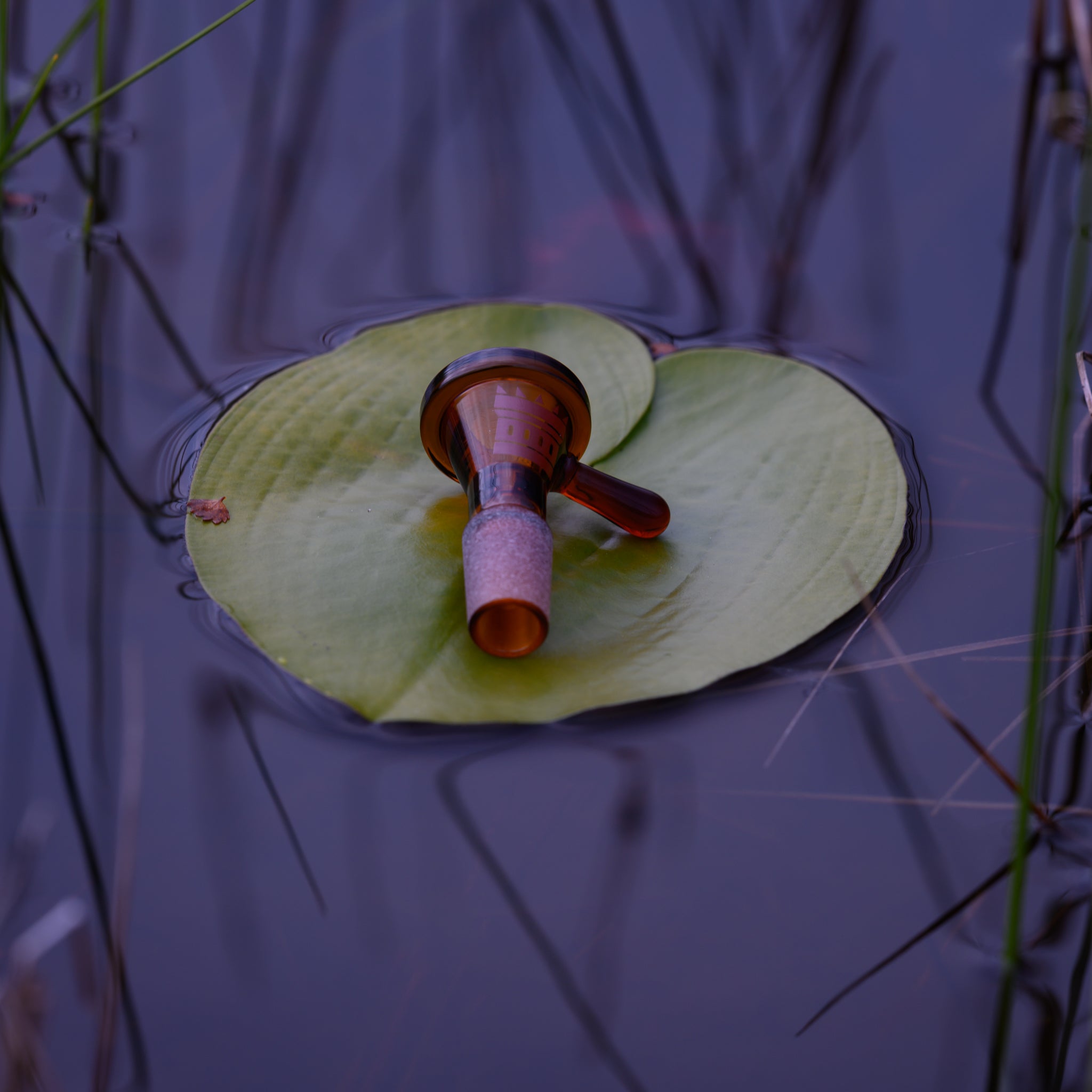A beautifully crafted amber glass bowl rests gently on a floating lily pad in a calm body of water. The smooth, rich hues of the glass contrast with the soft green tones of the lily pad, creating a serene and artistic composition. Reflections of reeds and plants in the water add depth to the image, enhancing the natural tranquility of the scene. The delicate balance of the bowl on the leaf highlights the craftsmanship and elegance of the glassware in harmony with nature.