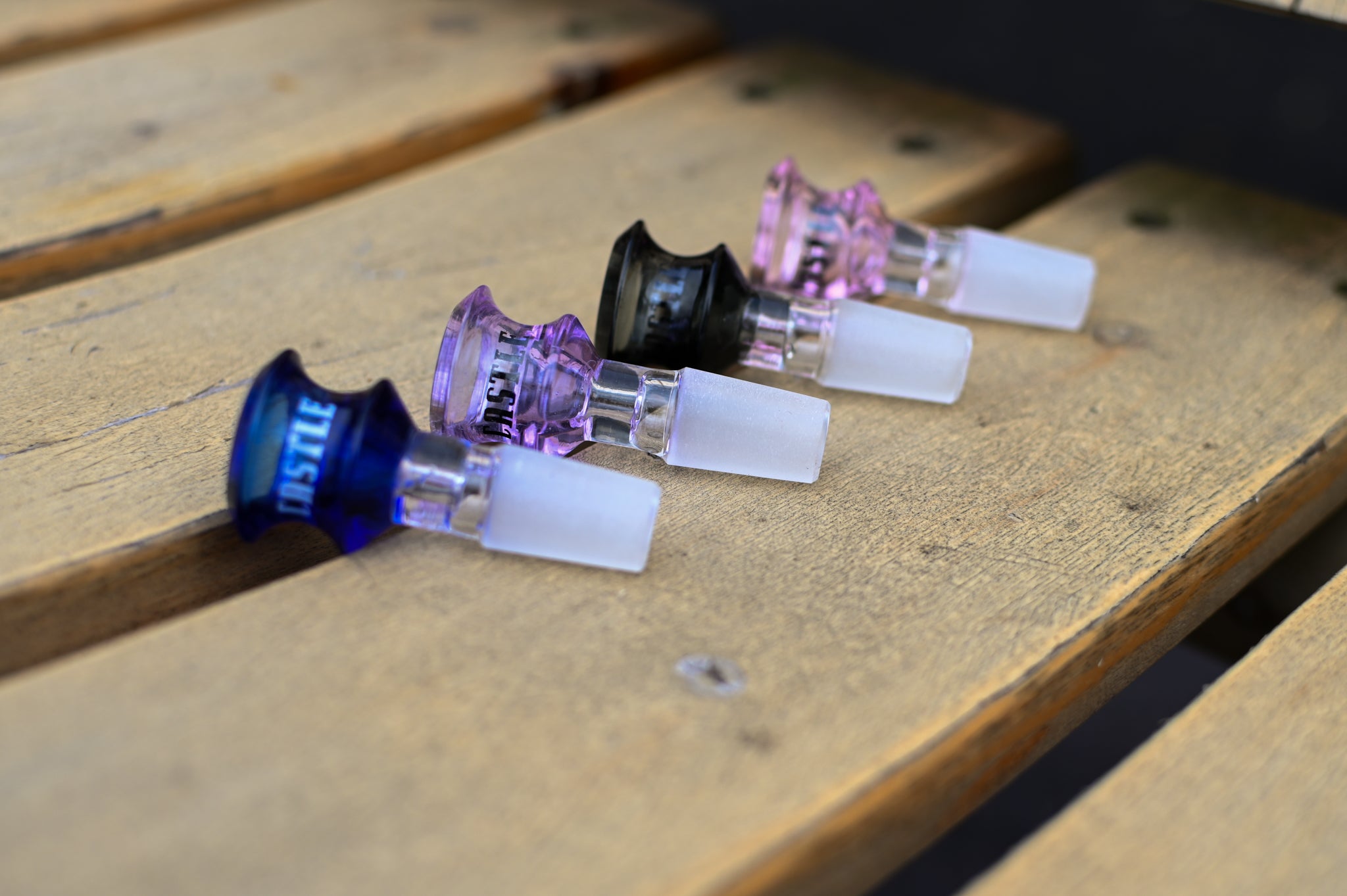 Close-up of four Castle-branded glass bowl attachments in different colors (blue, purple, black, and pink), each with a frosted male joint, lined up on a wooden bench.