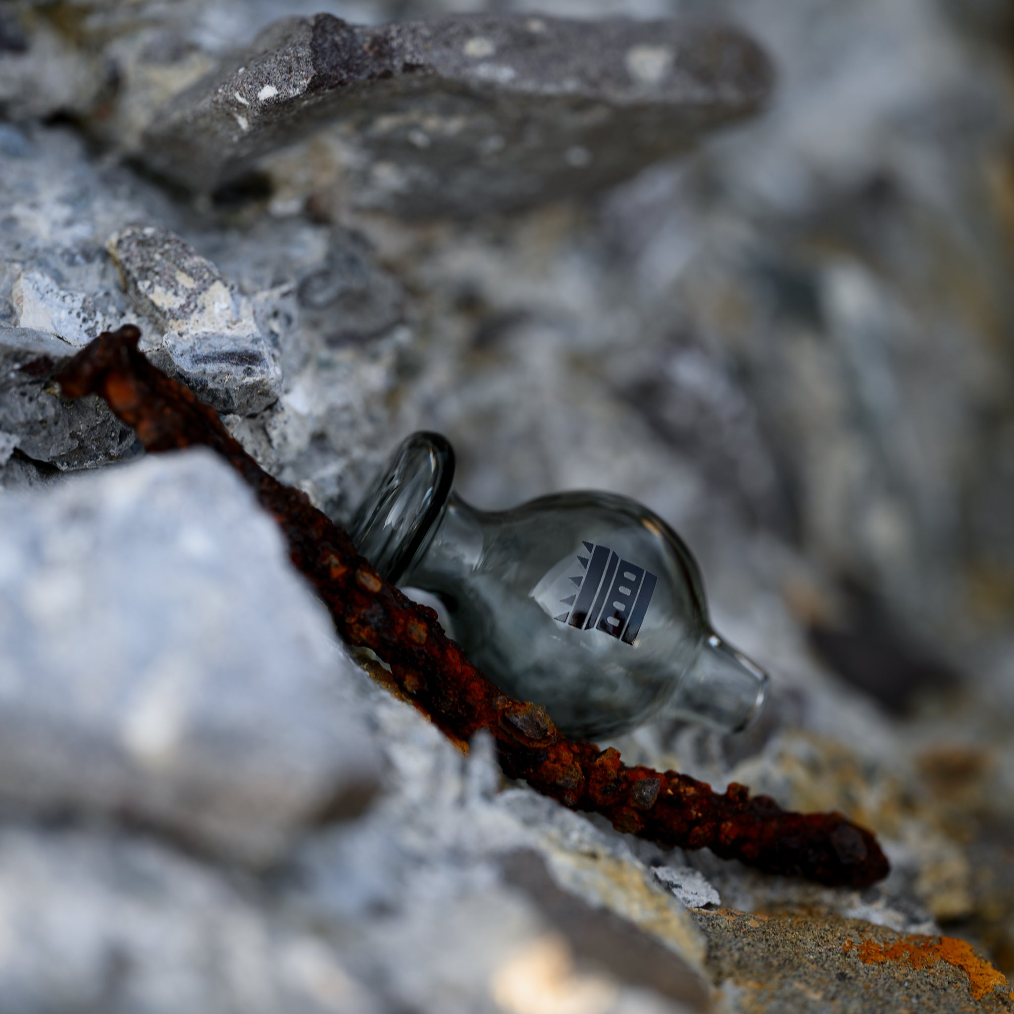 A smoky gray, Castle-branded glass carb cap with a rounded shape, resting among grey rocks beside a piece of rusted metal.