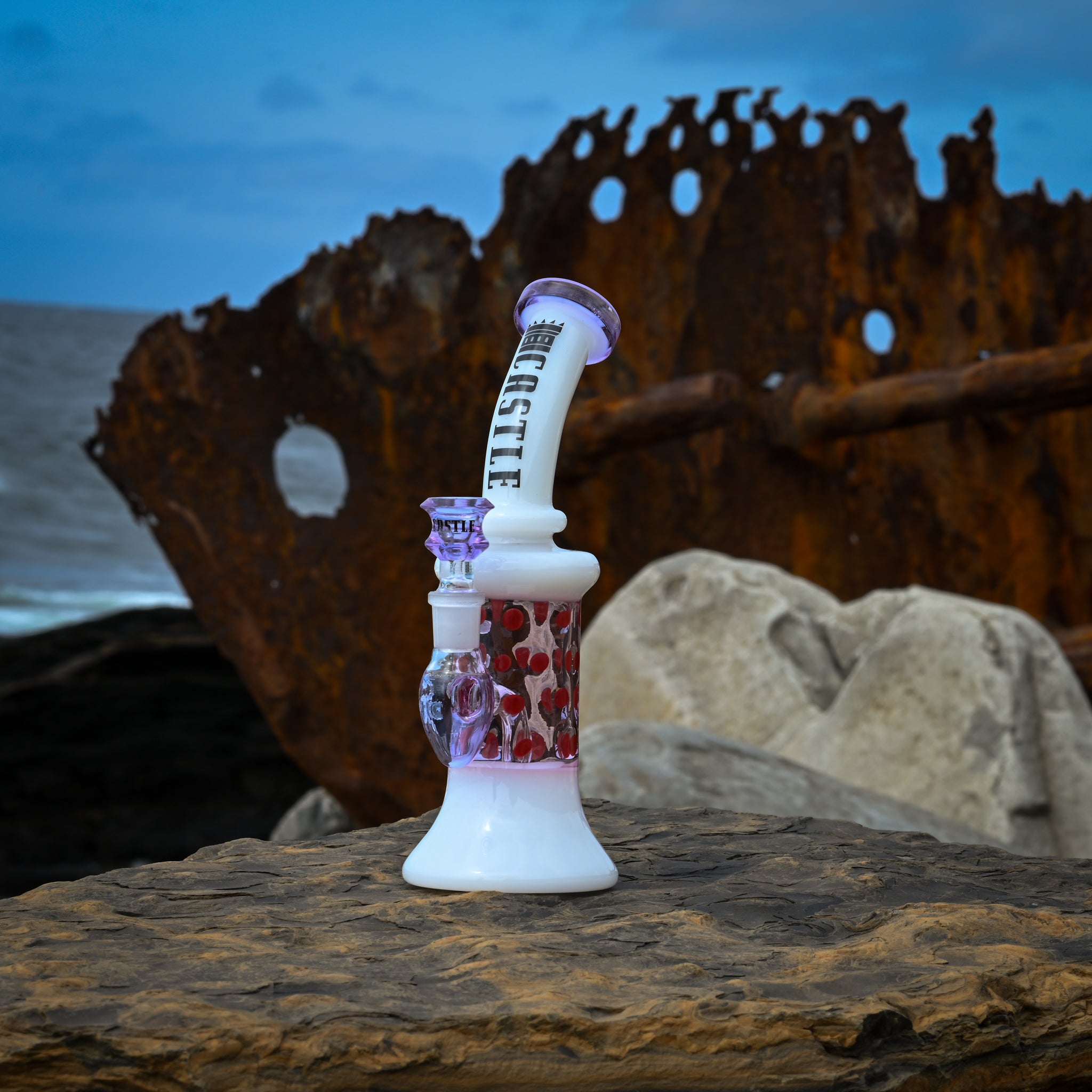 A white and pink Castle-branded glass dab rig with red polka dots, placed on a rock in front of a rusted shipwreck near the ocean.