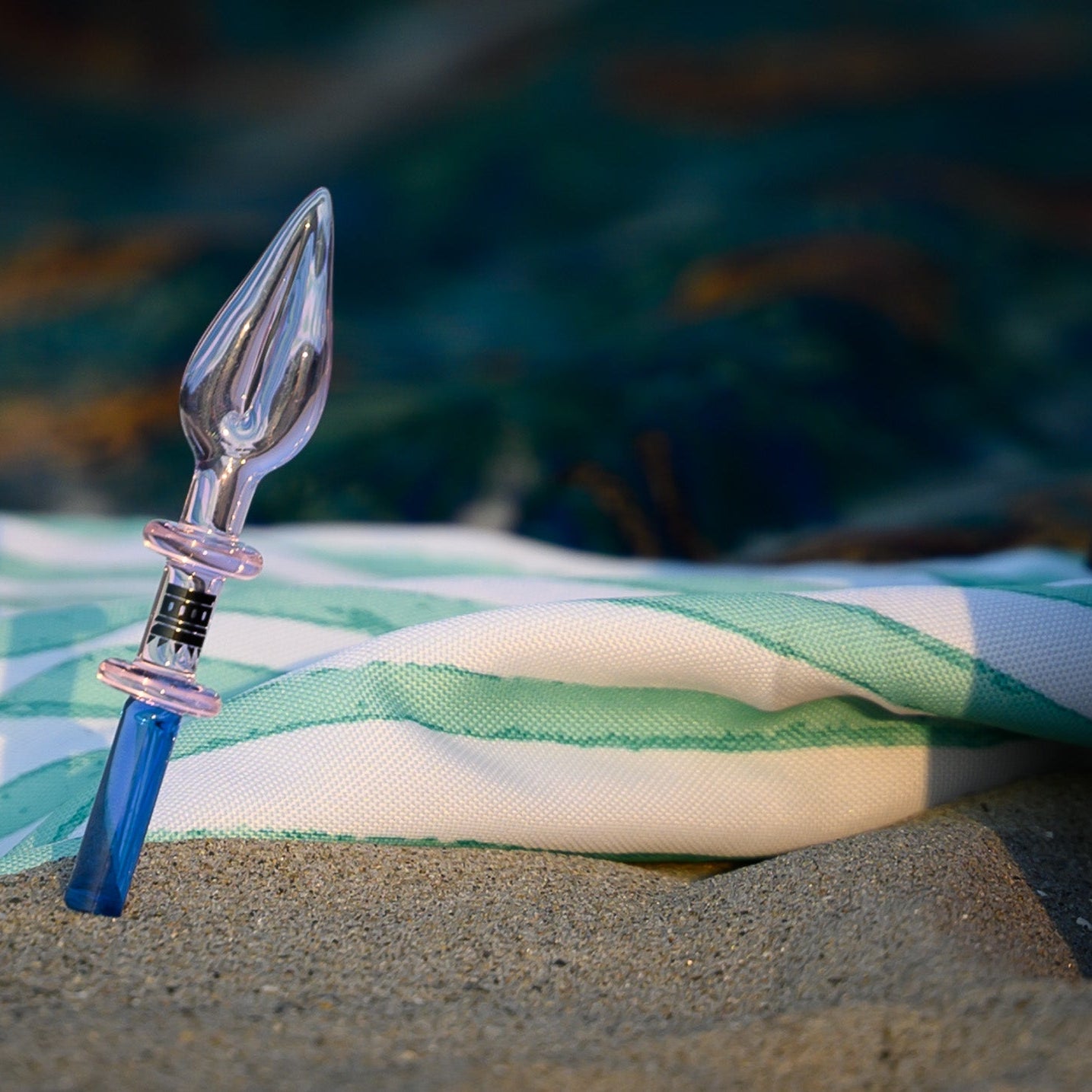 A beautifully crafted glass dab tool with a translucent pink and blue color gradient rests in the sand, partially propped against a green and white striped fabric. The tip of the tool features an elegant, pointed design, resembling a spear or flame, while the base transitions into a deep blue hue. In the blurred background, the ocean waves gently roll in, adding a serene and coastal ambiance to the scene. The composition highlights the tool’s artistry and functionality in a tranquil beach setting.