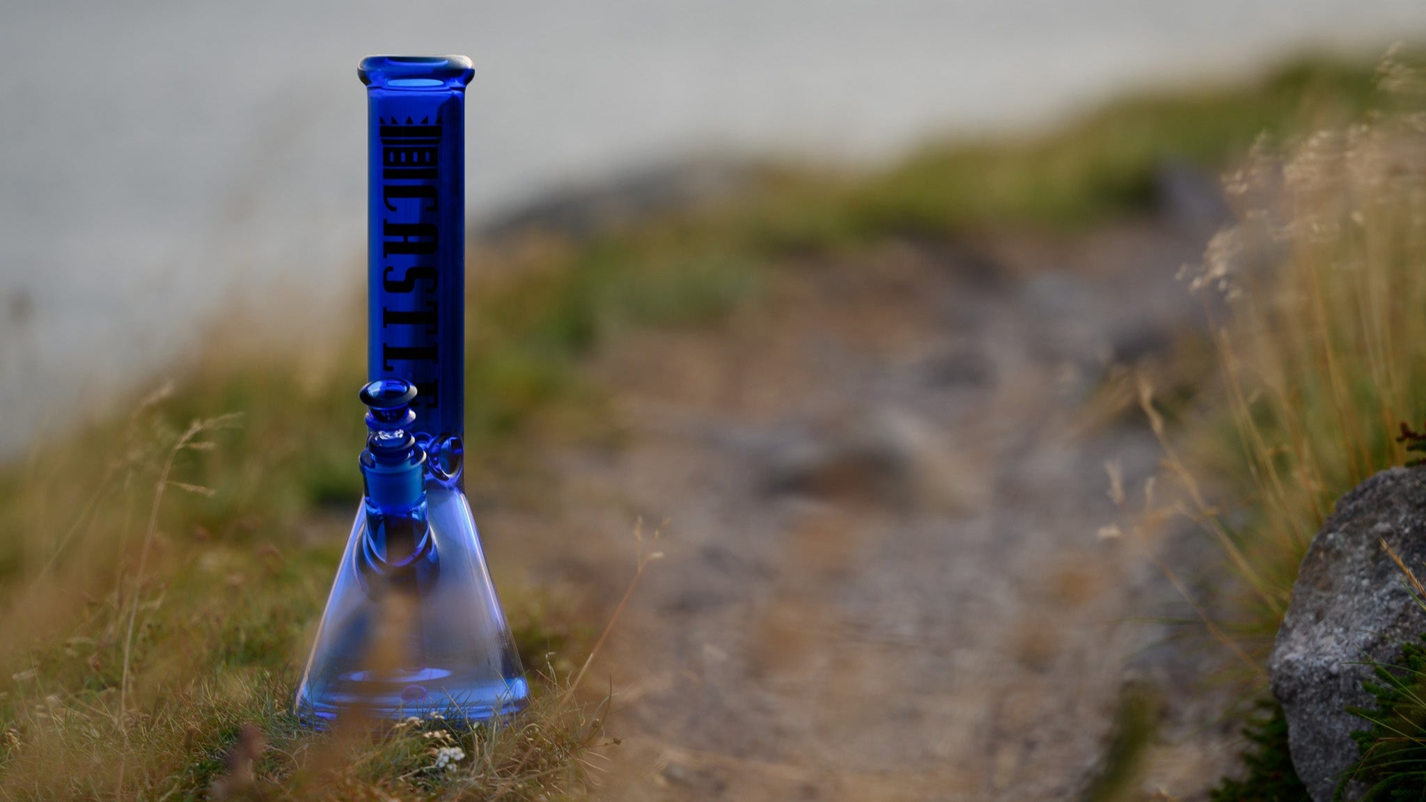 A cobalt-blue, beaker-style glass bong stands on a narrow dirt path overlooking a calm body of water, with golden grasses softly framing the foreground and “CASTLE” imprinted in bold lettering on the neck.