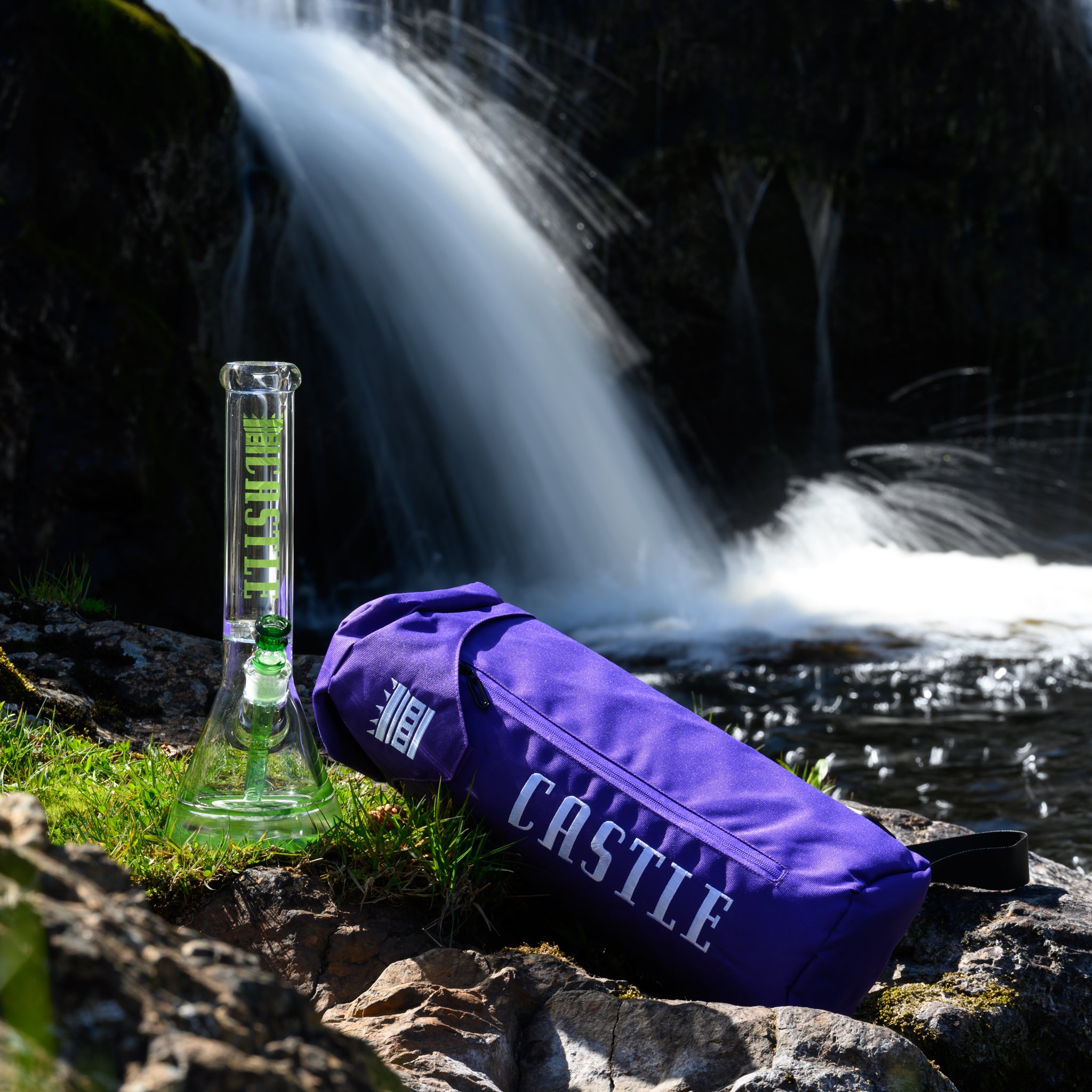 A clear glass water pipe with green highlights, branded ‘Castle,’ sitting on mossy rocks next to a purple Castle-branded carrying bag, with a cascading waterfall in the background.