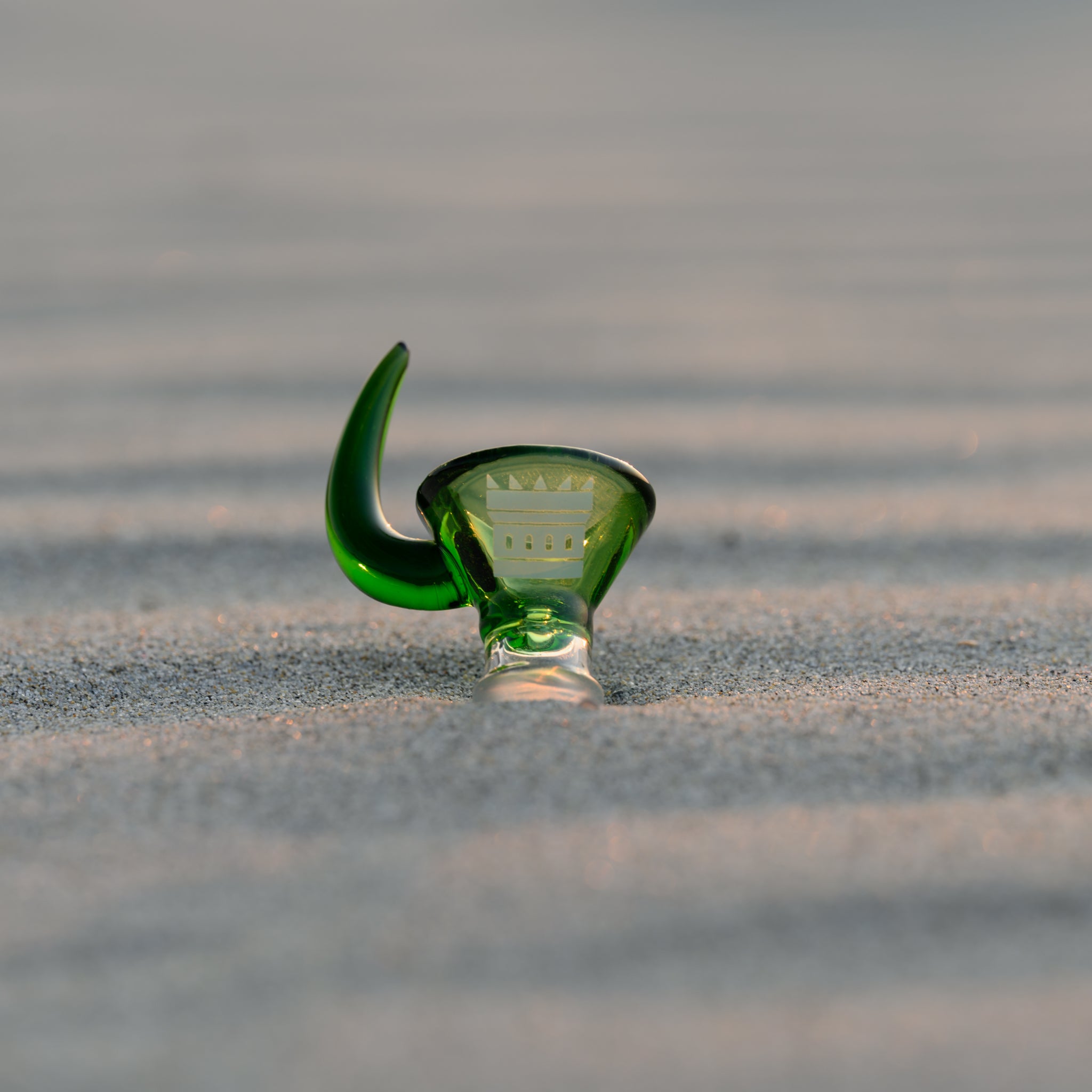 A green glass horn bowl with a castle logo etched inside, resting on rippled sand. The soft lighting highlights the smooth curves of the glass, creating a serene and minimalist composition.