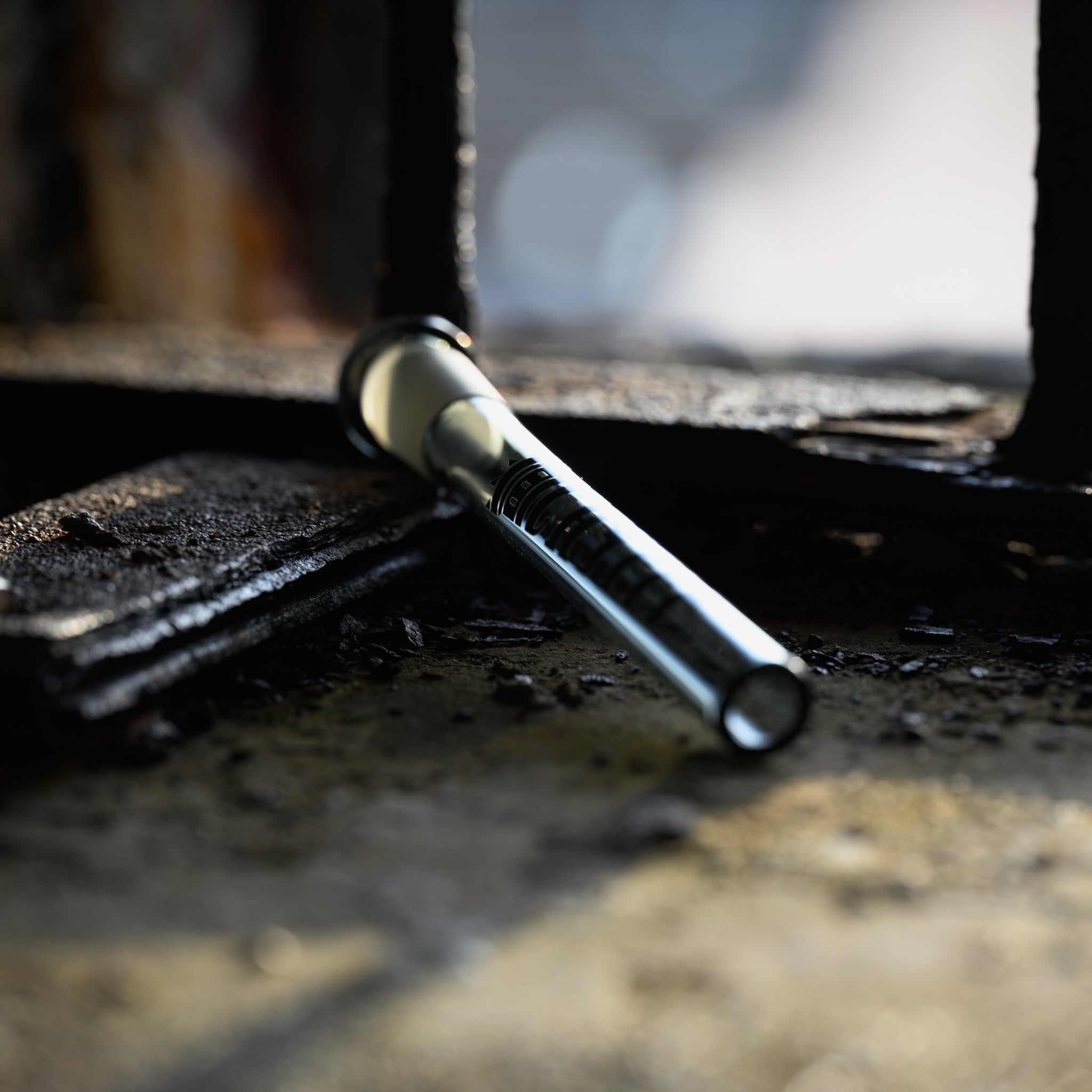 A close-up of a clear glass downstem, featuring faint black lettering near the top, lying diagonally on a rusted metal surface in a dimly lit setting. Soft light from the background highlights its cylindrical form against the industrial backdrop.