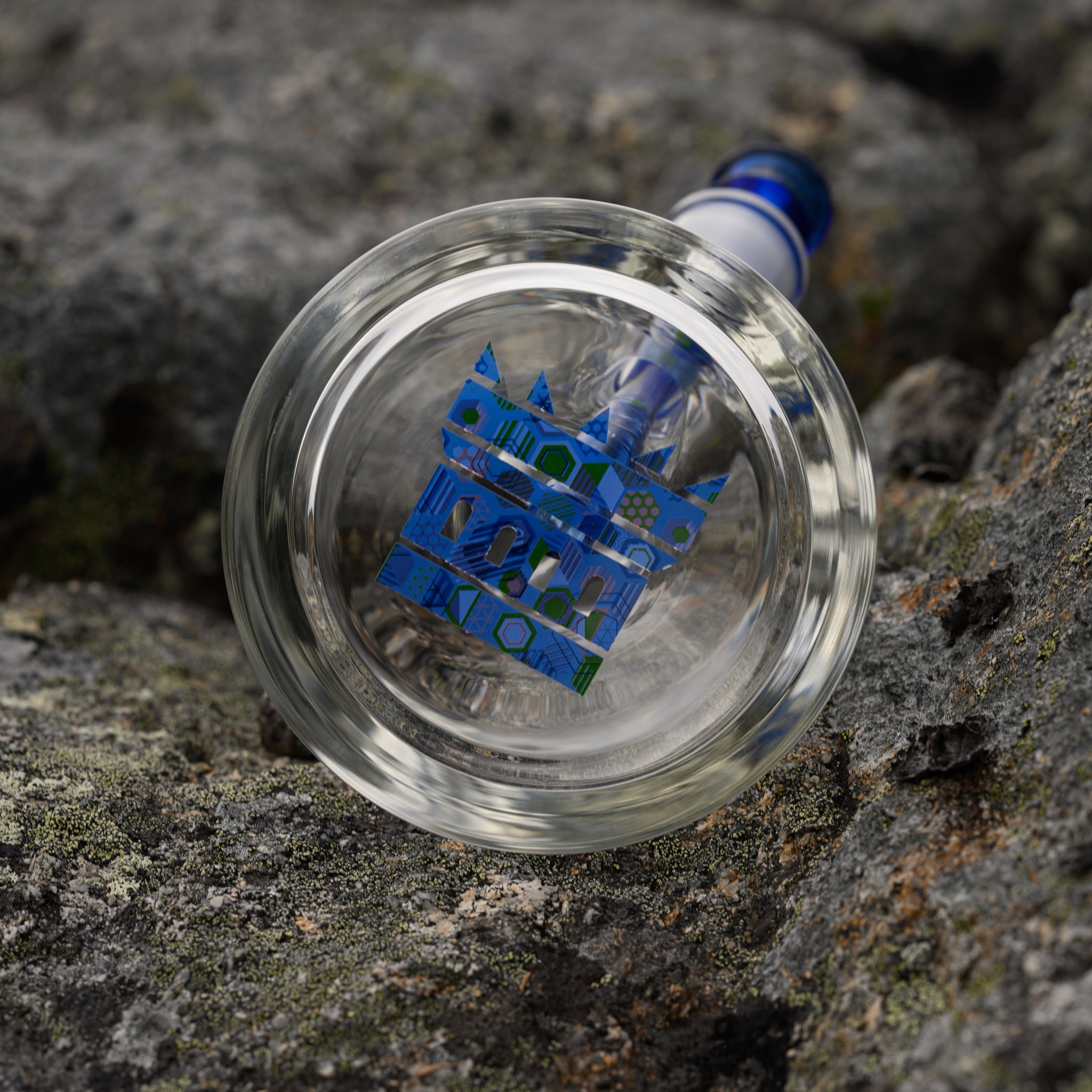 A close-up shot of the base of a high-quality glass bong, featuring an intricate blue castle logo at the center. The clear glass reveals the inner structure, including a blue-accented downstem, which matches the castle design. Resting on a rugged, textured rock surface, the image highlights the fine craftsmanship and durability of the piece. The natural setting contrasts with the sleek, modern glasswork, emphasizing the premium aesthetic of the bong.
