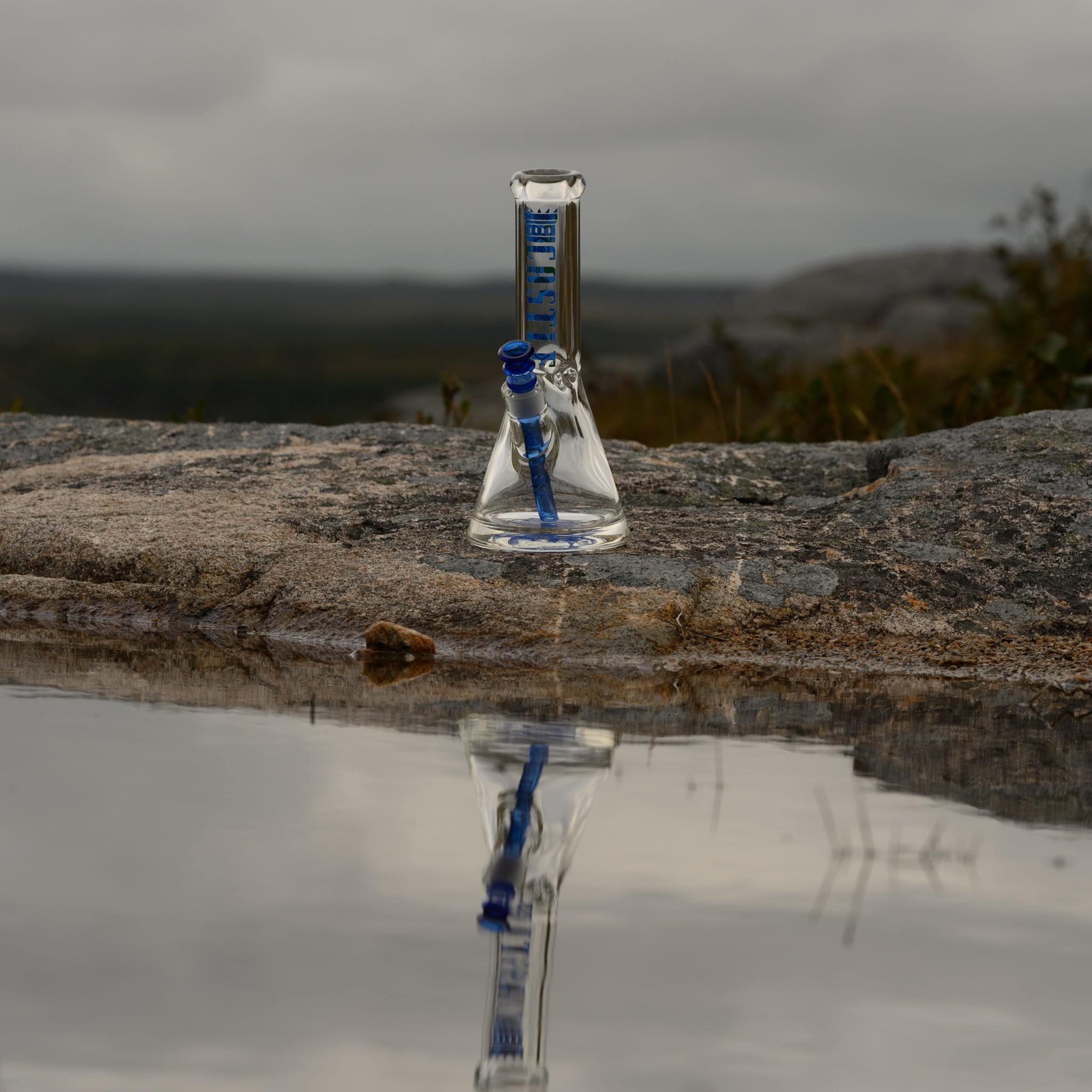 A sleek glass bong with bold blue accents from Castle Glassworks stands on a rocky surface near a still pool of water, reflecting its image. The beaker-style design features a blue-branded neck, matching downstem, and bowl, emphasizing its elegant craftsmanship. The overcast sky and rugged landscape create a moody, atmospheric backdrop, while the water’s reflection adds depth and symmetry to the composition, highlighting the bong’s premium aesthetic in a serene outdoor setting.