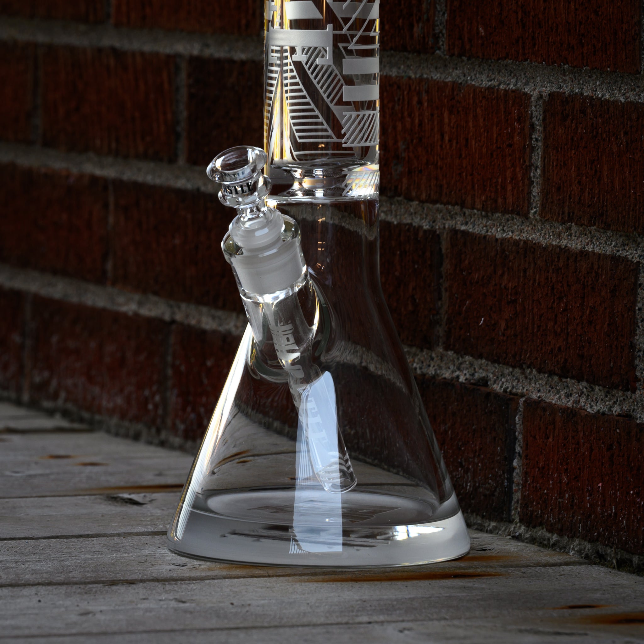 close-up view of a clear glass beaker bong with geometric designs etched onto its surface, positioned against a brick wall. The downstem and bowl are clearly visible, emphasizing the precision and clarity of the glass craftsmanship. The setting includes a wooden floor, adding to the rustic and urban feel of the scene. The combination of the modern, sleek design of the bong with the textured brick and wood backdrop creates an interesting contrast and visual appeal.