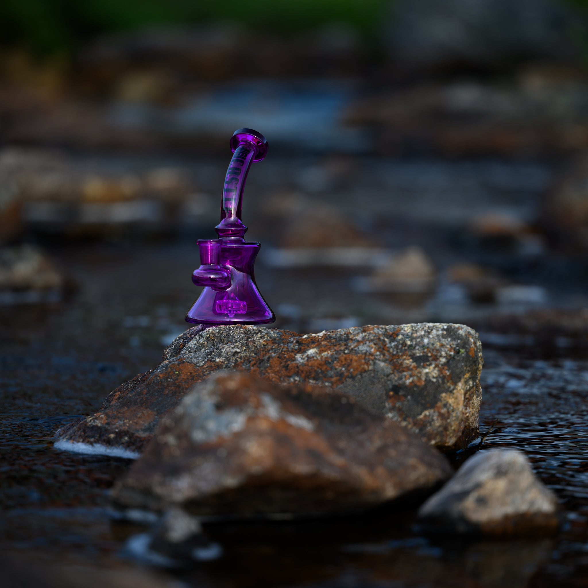 A vibrant purple mini dab rig with the "Castle" logo vertically displayed on the neck, positioned on a rock in the middle of a shallow river. The clear and colorful glass of the rig stands out against the natural backdrop of flowing water and stones. The surrounding greenery and the water's motion create a serene and picturesque scene, highlighting the contrast between the handcrafted glass and the organic setting.