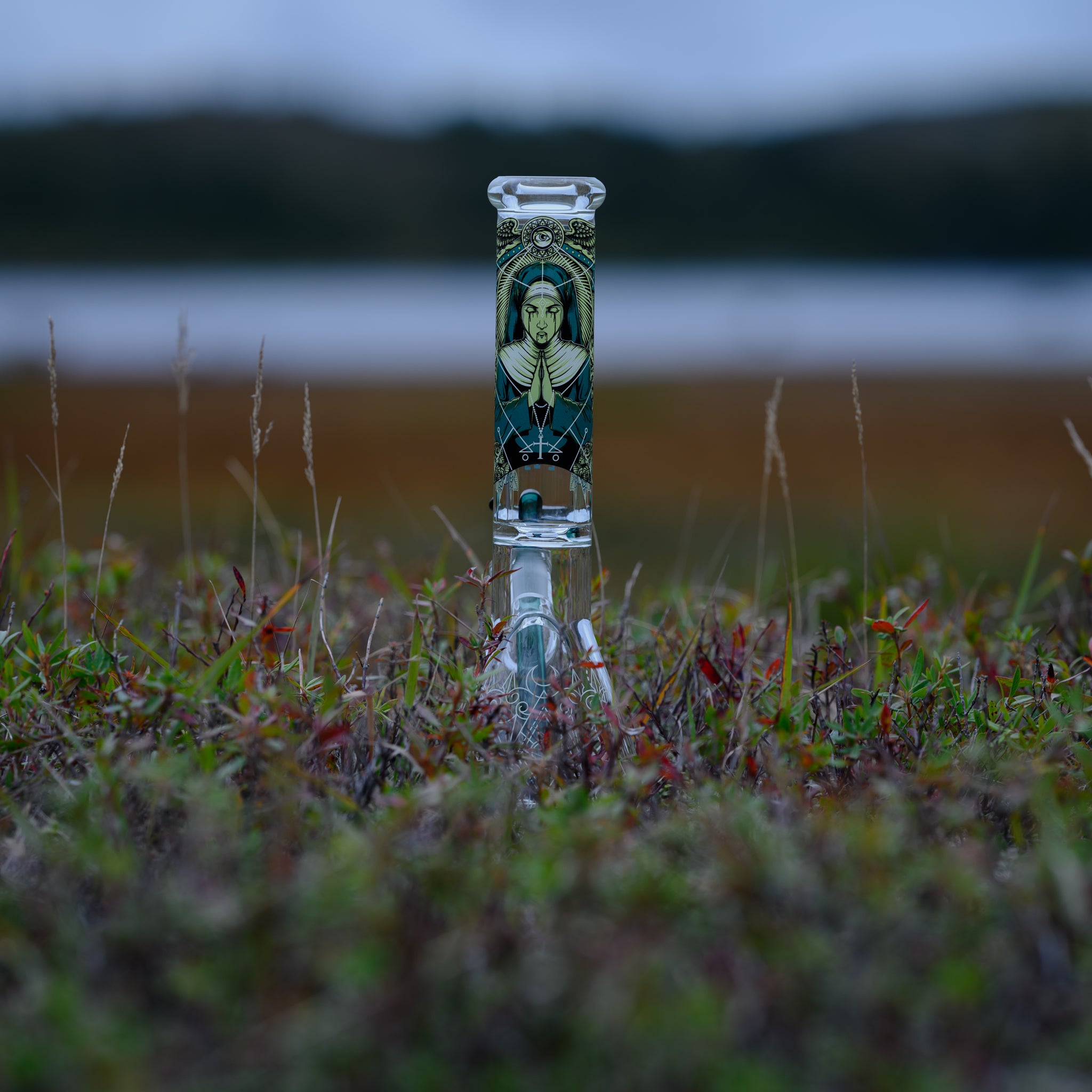 A glass bong with an intricate, gothic-inspired illustration on its neck stands amid a field of wild grass, with a blurred lake and rolling hills in the background. The artwork features a mysterious, regal woman adorned in ornate jewelry, set against a dark, geometric backdrop. The bong's clear glass base blends seamlessly with the natural surroundings, while the detailed design on the neck draws attention to its unique aesthetic. The moody, overcast lighting enhances the dramatic appeal of the bong.