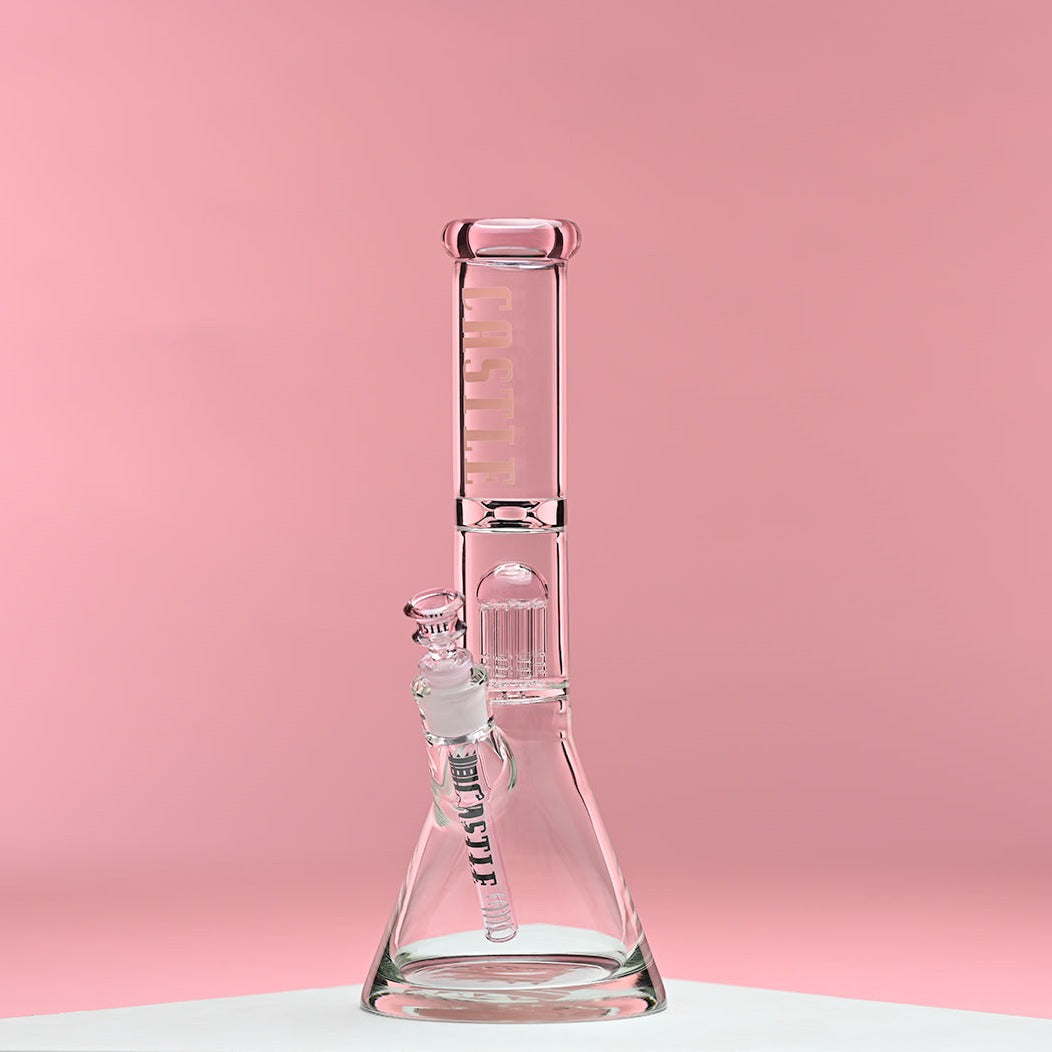 A glass beaker bong with pink accents and the Castle Glassworks logo on the neck. The bong features an eight-arm percolator and is set against a soft pink background, highlighting its elegant design and intricate details. The overall aesthetic is clean and modern, emphasizing the craftsmanship and quality of the piece.