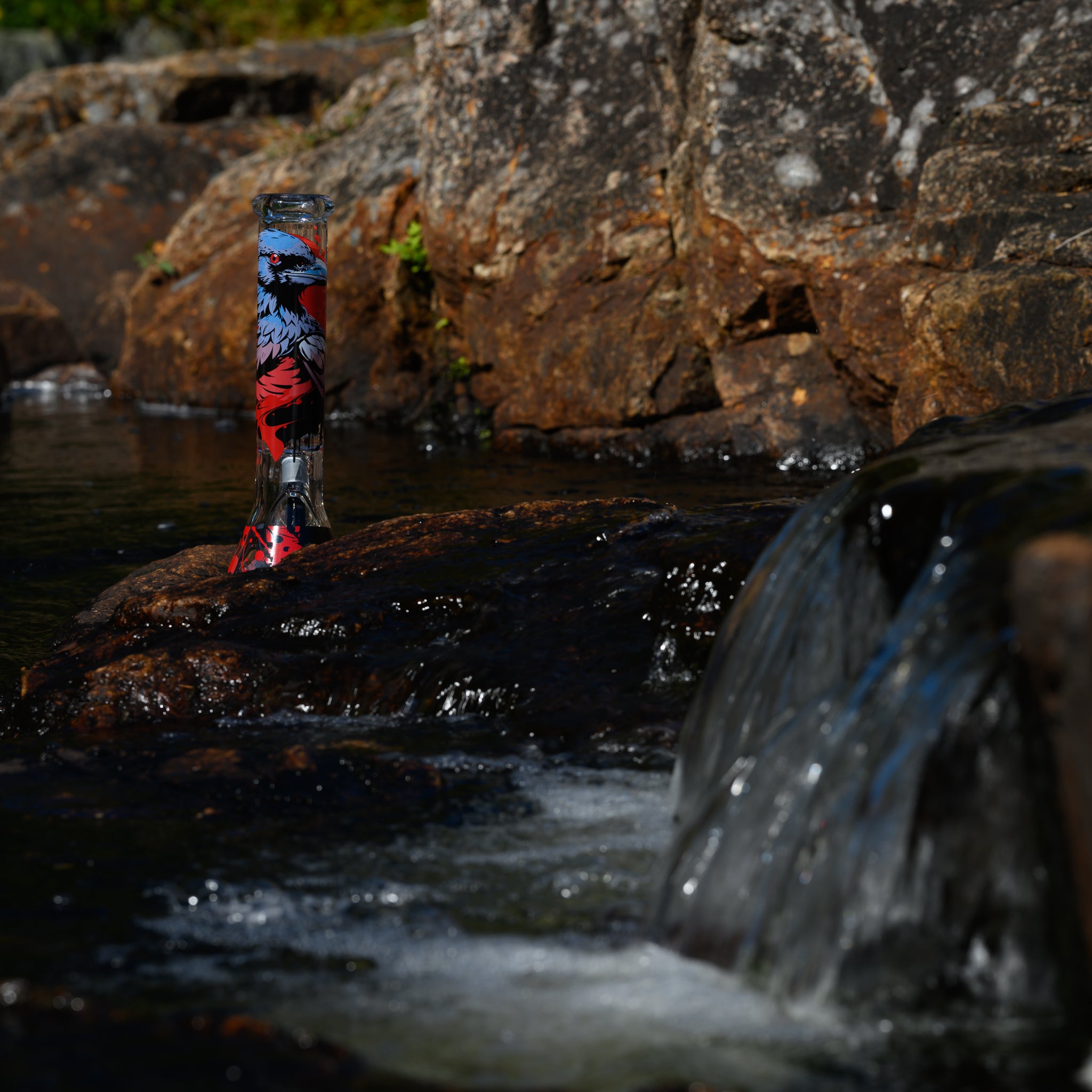 A glass bong with bold red, black, and white artwork stands on a wet rock beside a gently flowing waterfall. The intricate design wraps around the neck, creating a striking contrast against the clear glass base. The natural setting, with cascading water and rugged stone formations, enhances the adventurous and untamed aesthetic of the piece. Sunlight casts dramatic shadows, highlighting the craftsmanship and durability of the bong in this serene outdoor environment.
