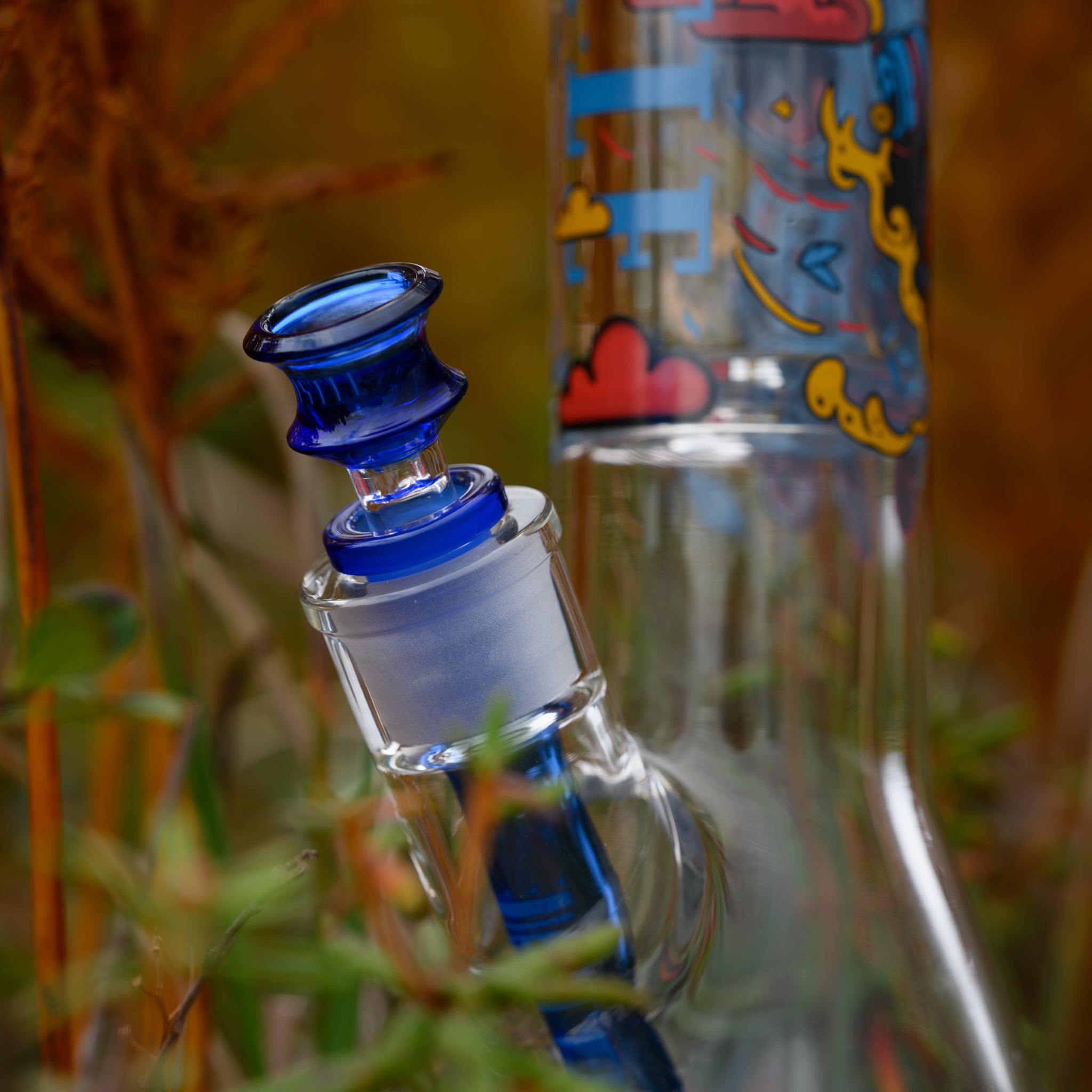 A close-up of a glass bong highlighting its vibrant blue bowl and matching downstem, set against a backdrop of wild greenery. The bowl features a rich cobalt hue with a multi-tiered design, complementing the frosted glass joint. The neck of the bong showcases playful, colorful artwork with abstract elements, clouds, and dynamic lines. The intricate craftsmanship of the glass and the detailed design contrast beautifully with the natural environment, emphasizing both function and artistic expression.
