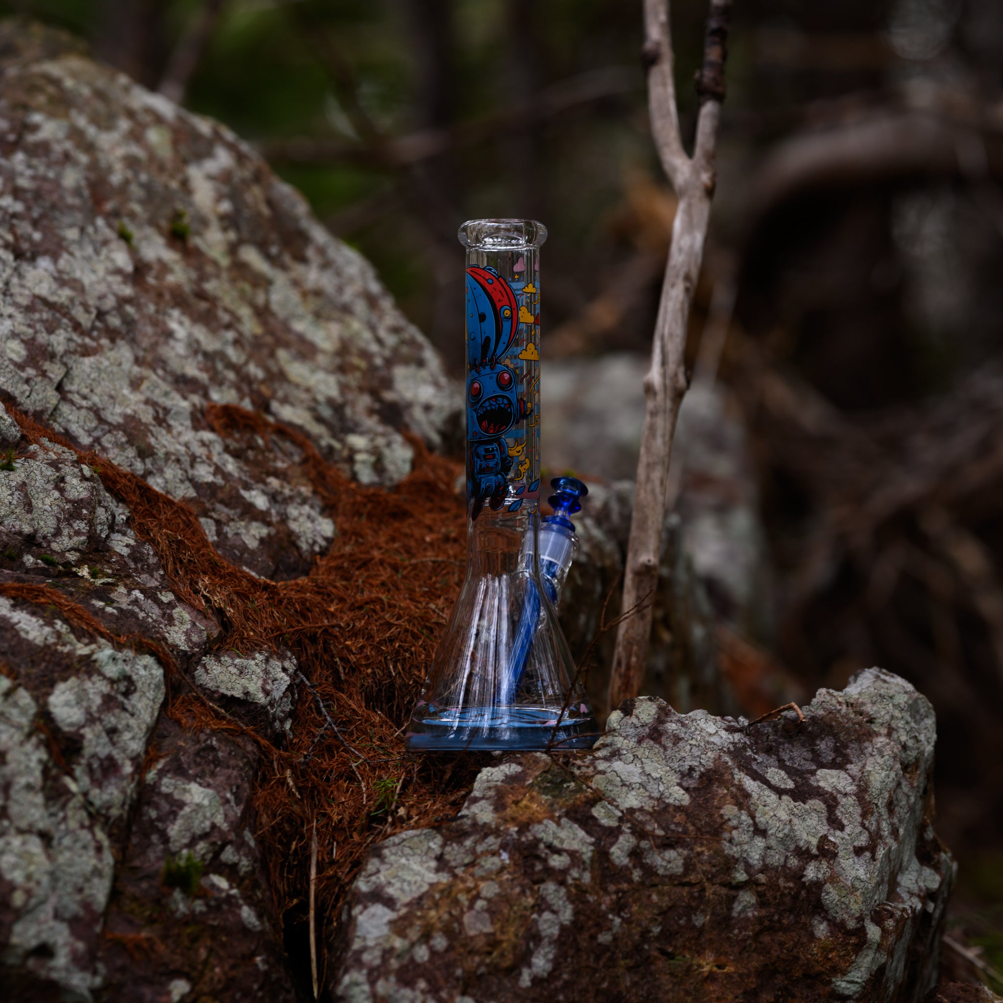 A clear beaker-style glass bong featuring cartoon robot illustrations and a matching blue downstem is perched on a rocky surface covered with lichen and rust-colored moss. The textured rock and surrounding branches frame the piece against a forest backdrop.