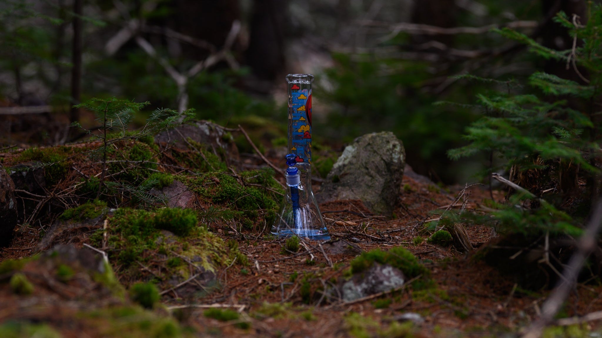 A tall, clear beaker-style glass bong adorned with colorful robot graphics and a matching blue downstem stands on a moss- and pine-needle-covered forest floor. It’s surrounded by rocks and evergreen trees, with a soft, natural light illuminating the glass against the muted woodland backdrop.