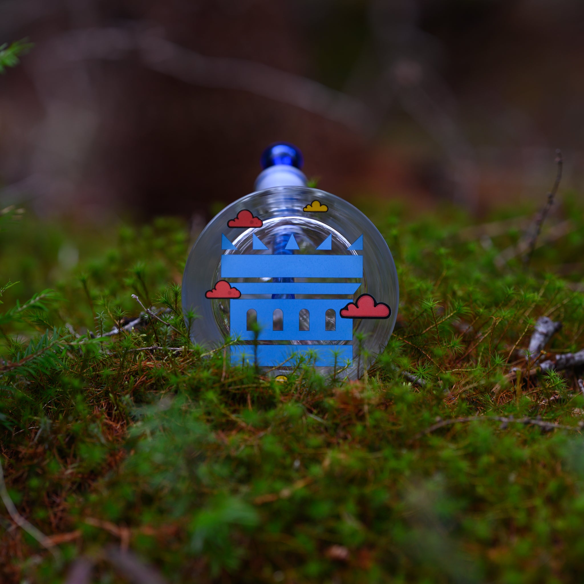 A glass bong decorated with a stylized blue castle design and small colorful clouds sits on a lush bed of green moss.