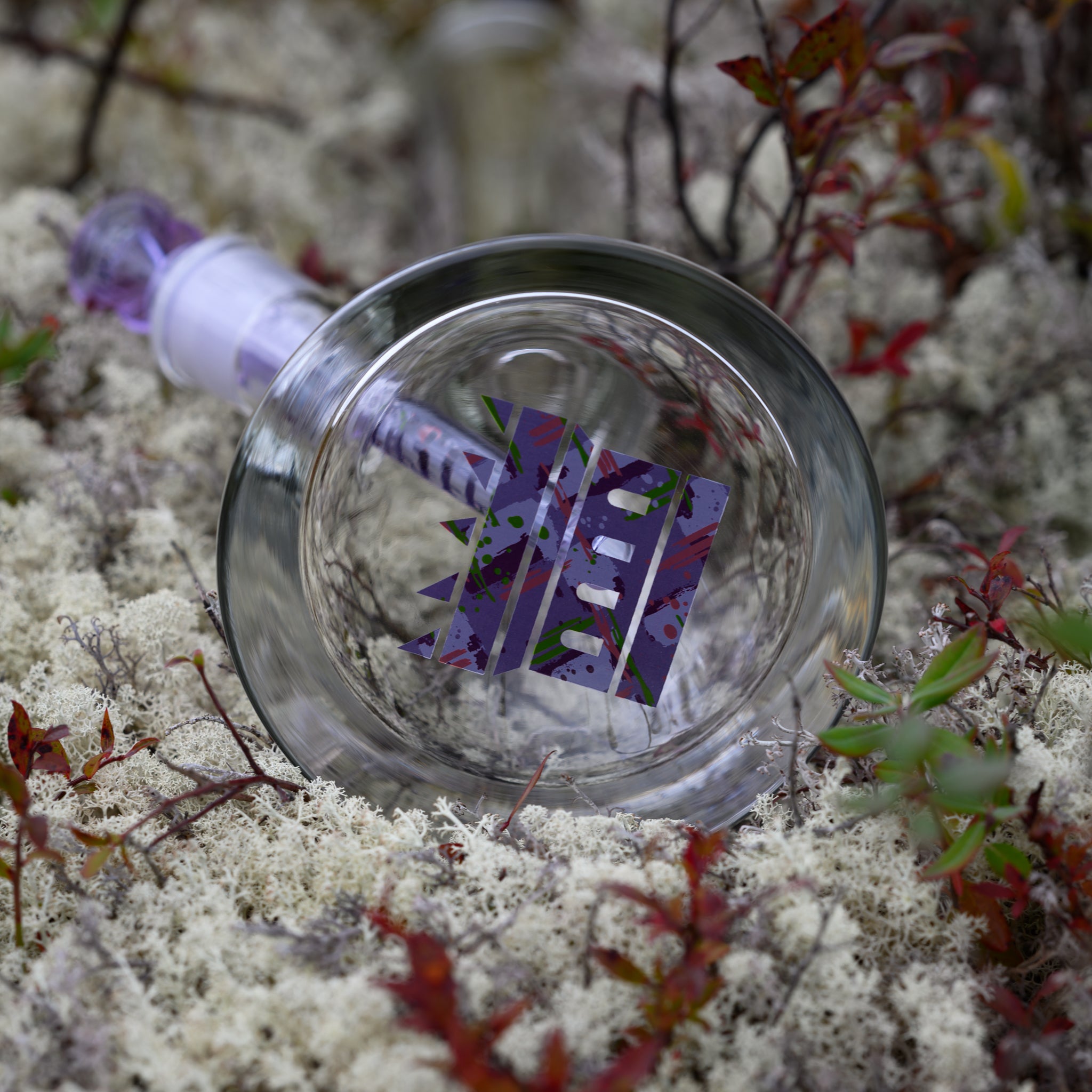 A close-up of the base of a glass bong resting on a bed of soft white moss, surrounded by delicate red and green foliage. The bottom features a striking purple geometric "CASTLE" logo, which contrasts beautifully against the transparent glass. The matching purple-accented downstem and bowl add cohesion to the design. The organic textures of the moss and plants complement the smooth glass, creating a visually balanced composition that highlights both craftsmanship and nature’s beauty.