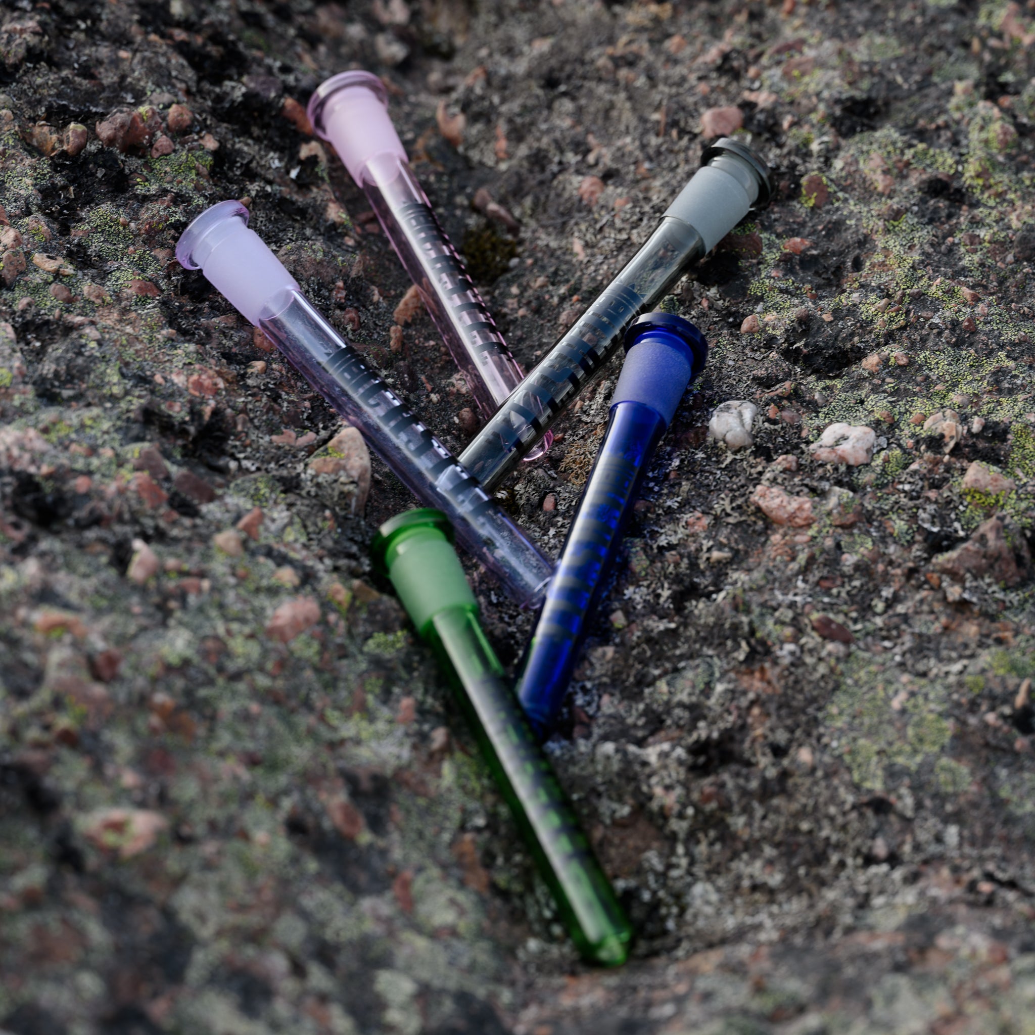 A collection of colorful glass downstems with the Castle Glassworks Logo are arranged in a fan-like pattern on a rocky, moss-covered surface. The vibrant colors of the downstems contrast with the rough texture and earthy tones of the rocks, highlighting their sleek and polished design. The scene is naturally lit, emphasizing the downstems' smooth finish and intricate details.