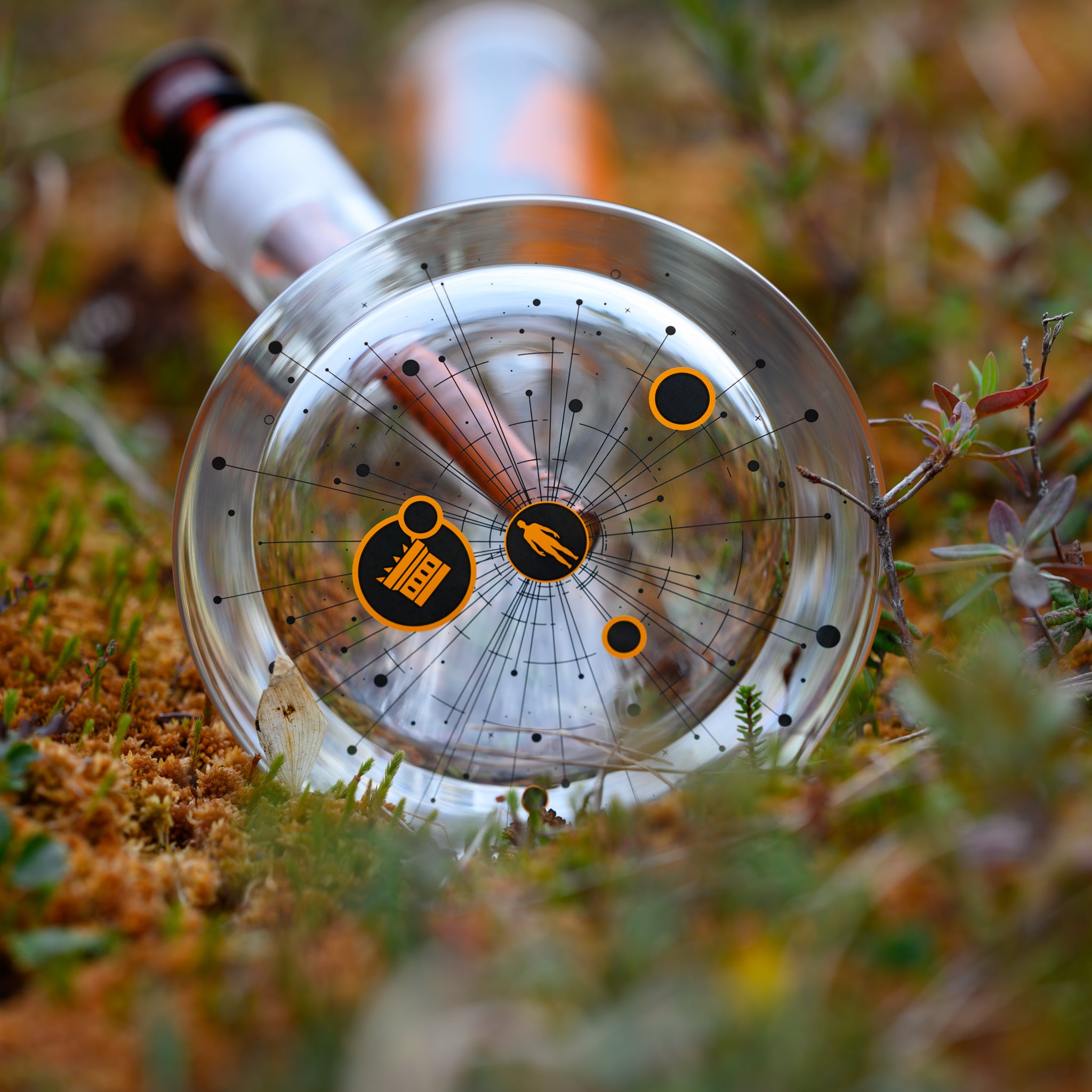 A close-up of the base of a glass bong showcases an intricate cosmic-inspired design etched in black and orange. The pattern features orbiting lines, planetary symbols, and small icons, including a figure being abducted and the Castle Glassworks logo, reinforcing the UFO theme. The transparency of the glass highlights the amber-hued downstem and bowl, creating a futuristic aesthetic. Resting on a bed of moss and wild vegetation.