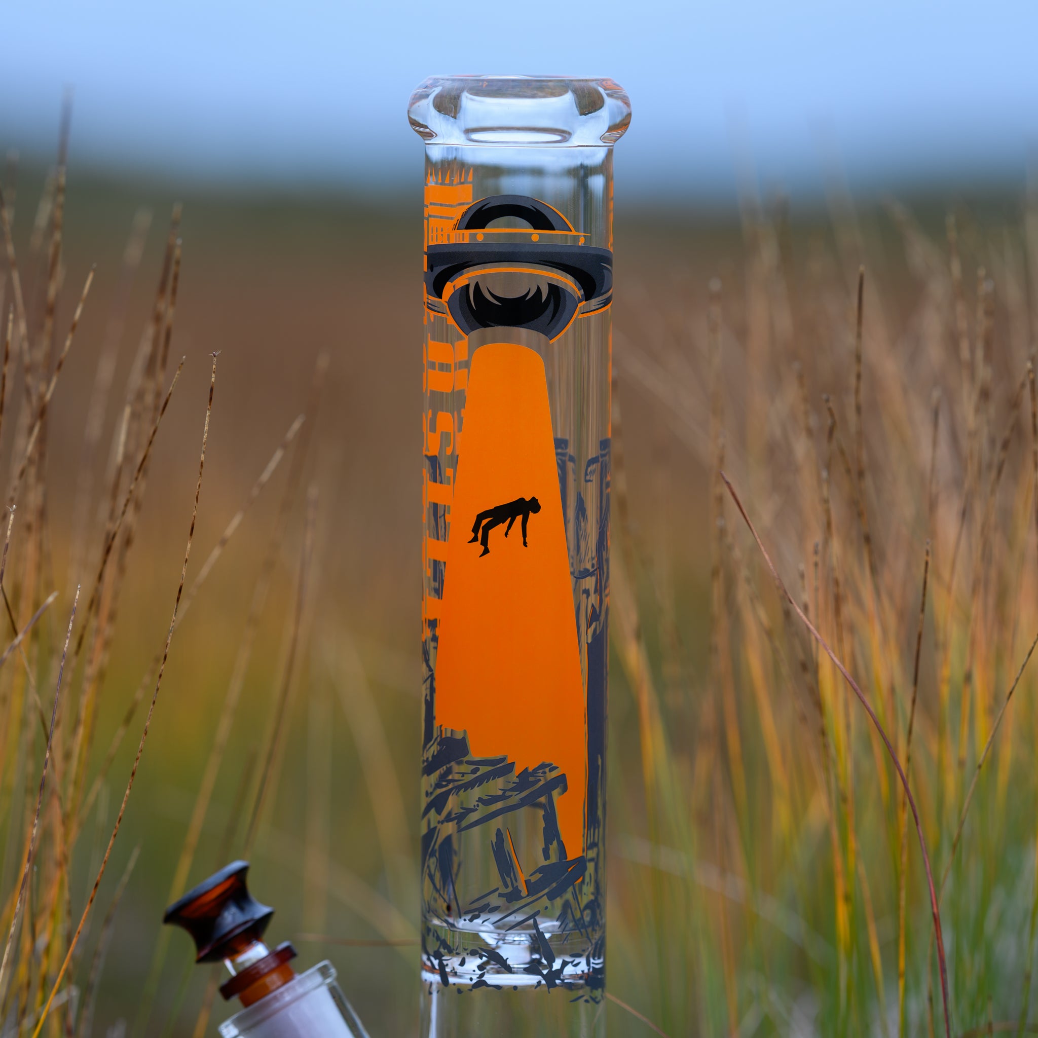 A striking glass bong featuring an eye-catching UFO abduction design stands tall among wild prairie grass. The artwork on the neck depicts a dark spaceship beaming up a silhouetted figure with a bold orange glow, creating a dramatic contrast against the clear glass. The "CASTLE" branding is subtly integrated into the design. Below, a rich amber-hued glass bowl complements the extraterrestrial theme.