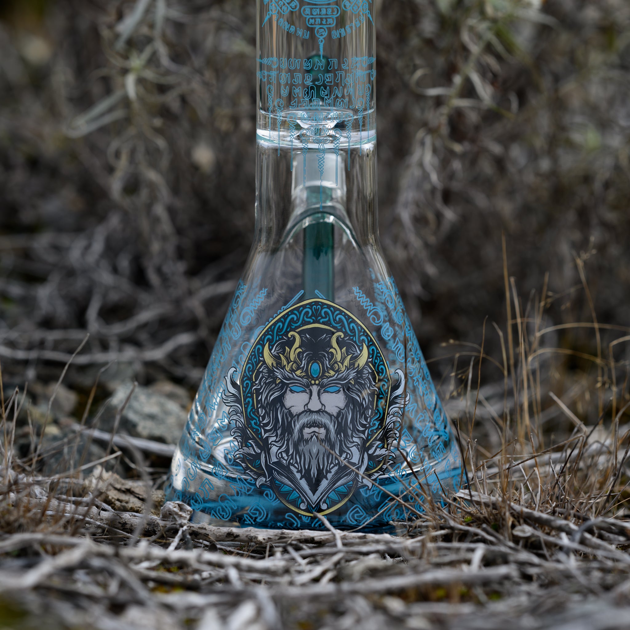 Close-up of the beaker base of a clear glass bong, decorated with an ornate Norse-inspired illustration of a bearded figure in a horned crown, surrounded by teal runic patterns. The piece rests among dried grasses, emphasizing the detailed artwork against the natural outdoor setting.