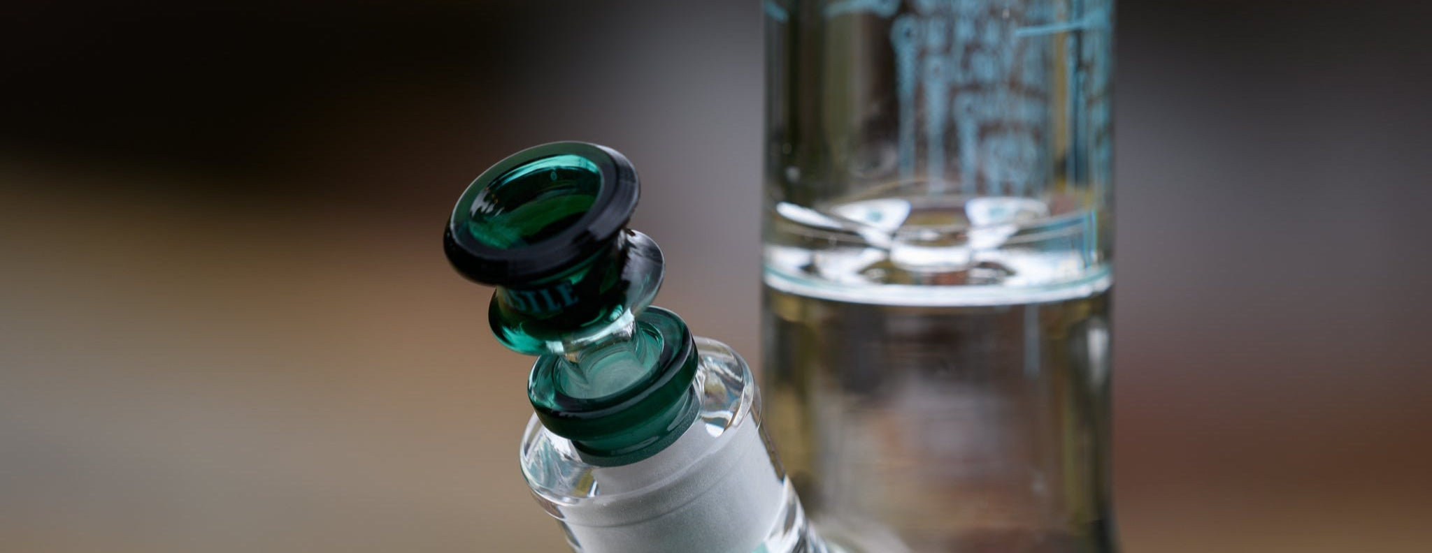 Close-up of a clear glass beaker-style water pipe adorned with teal artwork. The matching teal glass bowl piece is inserted into the downstem, and the background is softly blurred.