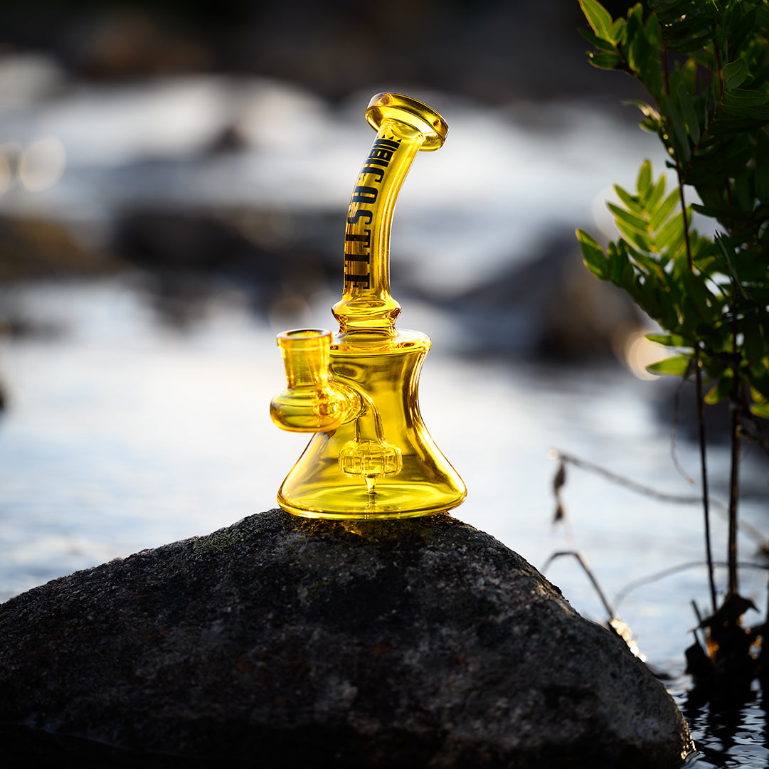 A vibrant yellow glass mini dab rig sits on a smooth rock by the water's edge. The rig features a curved neck with the "CASTLE" logo in bold black lettering. Sunlight reflects off the water and the rig, creating a bright and striking scene. Green foliage frames the image on the right, adding a natural element to the setting.