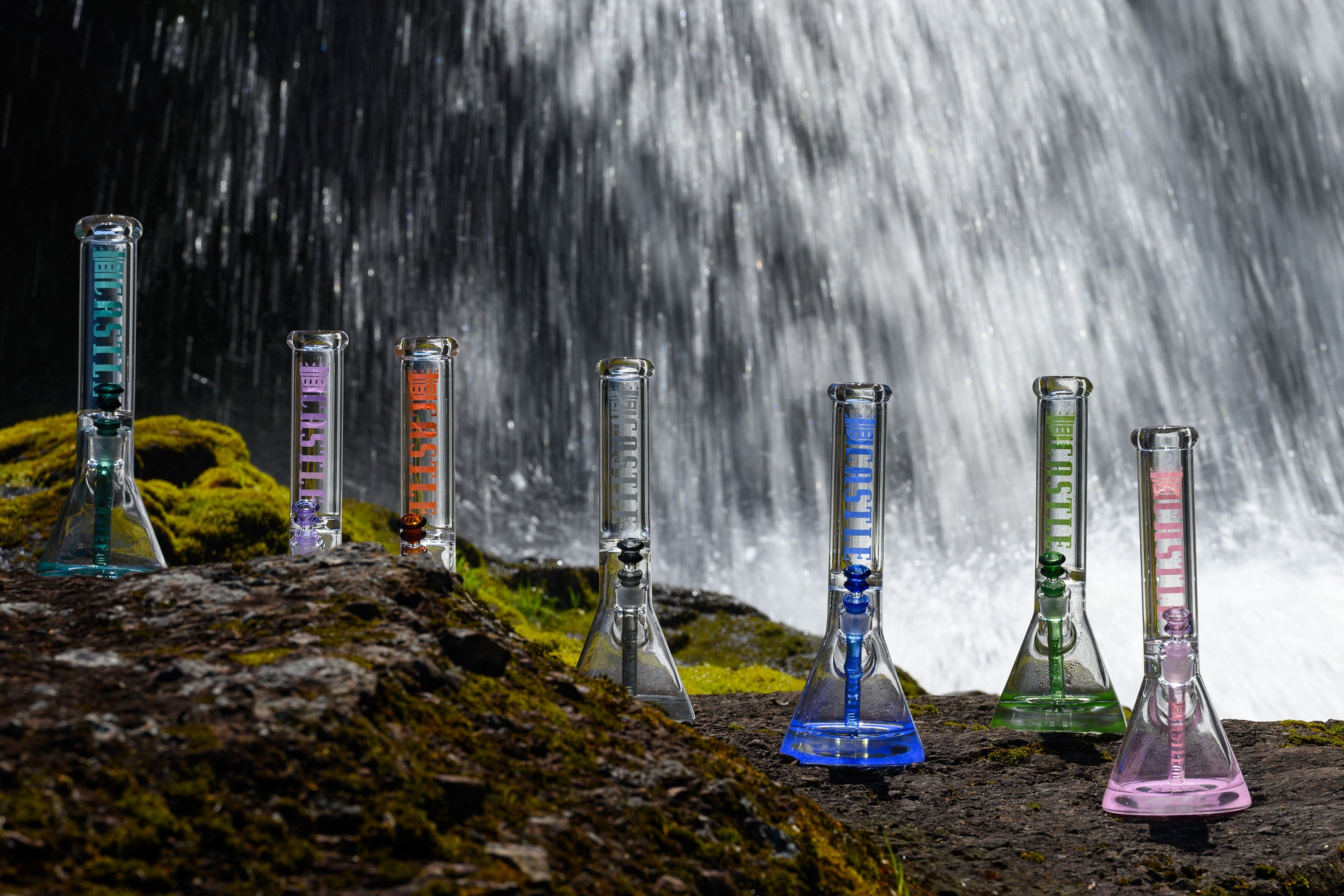 A collection of beaker bongs, each with colorful accents, on moss-covered rocks in front of a cascading waterfall.