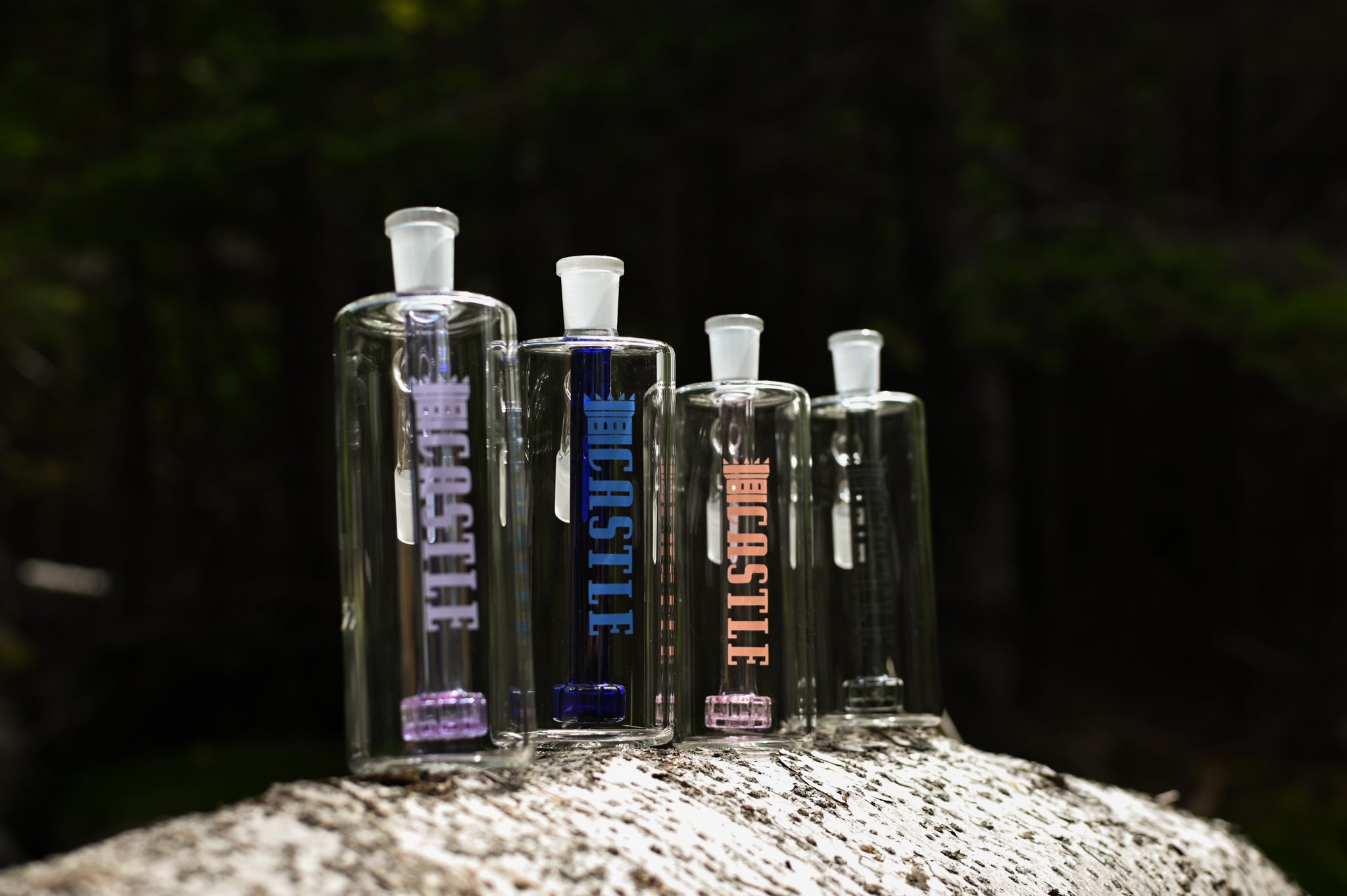 Four clear glass Castle-branded ash catchers with differently colored percolators (purple, blue, pink, and grey), standing upright on a pale rock in a forested background.