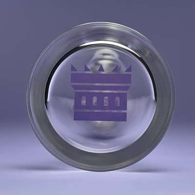 Close-up of the bottom base of a glass bong featuring a purple castle logo. The logo, centrally placed, stands out against the clear glass, set against a solid purple background that complements the design.
