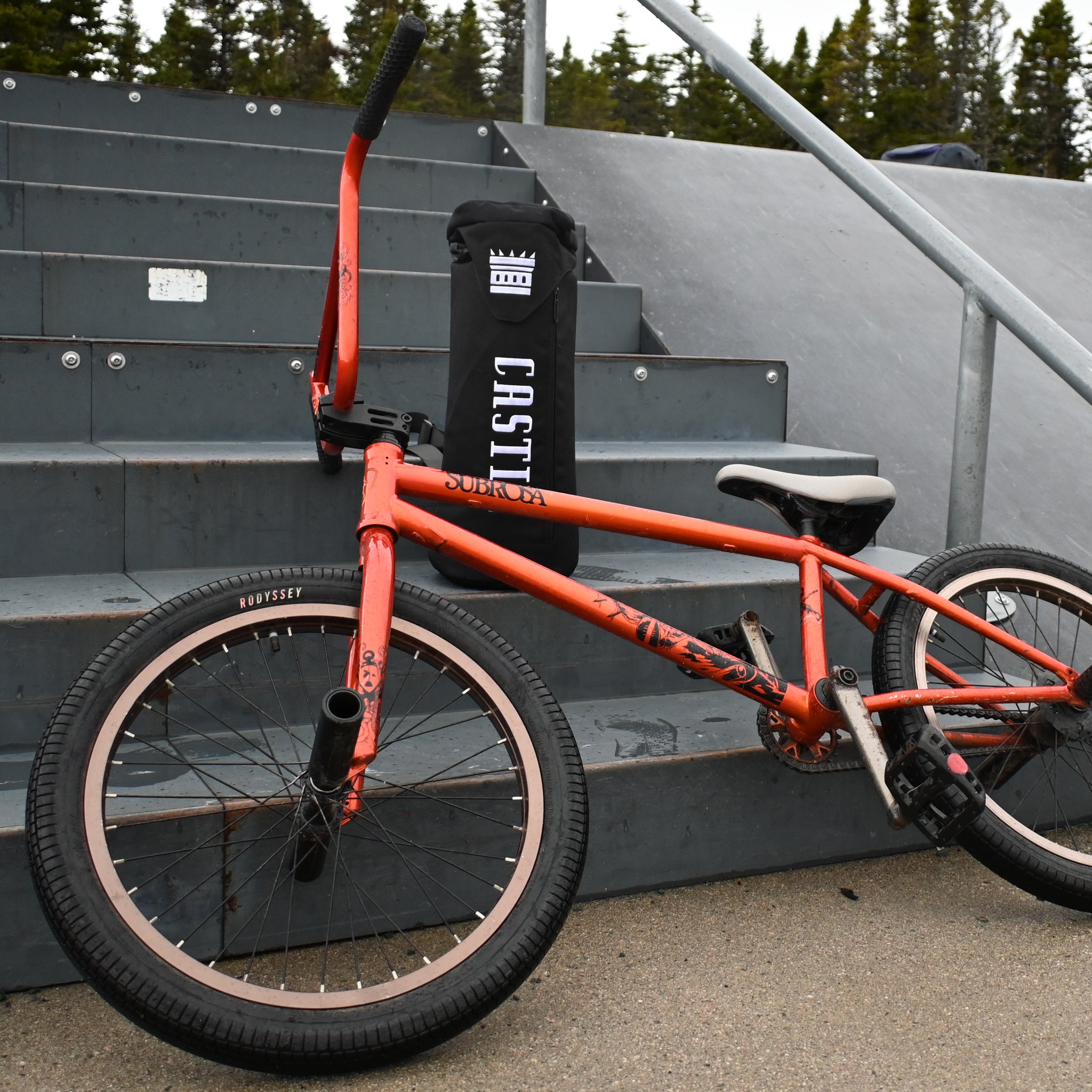An orange BMX bike resting against metal steps at a skatepark, with a black Castle-branded bong bag standing upright on a step behind it.