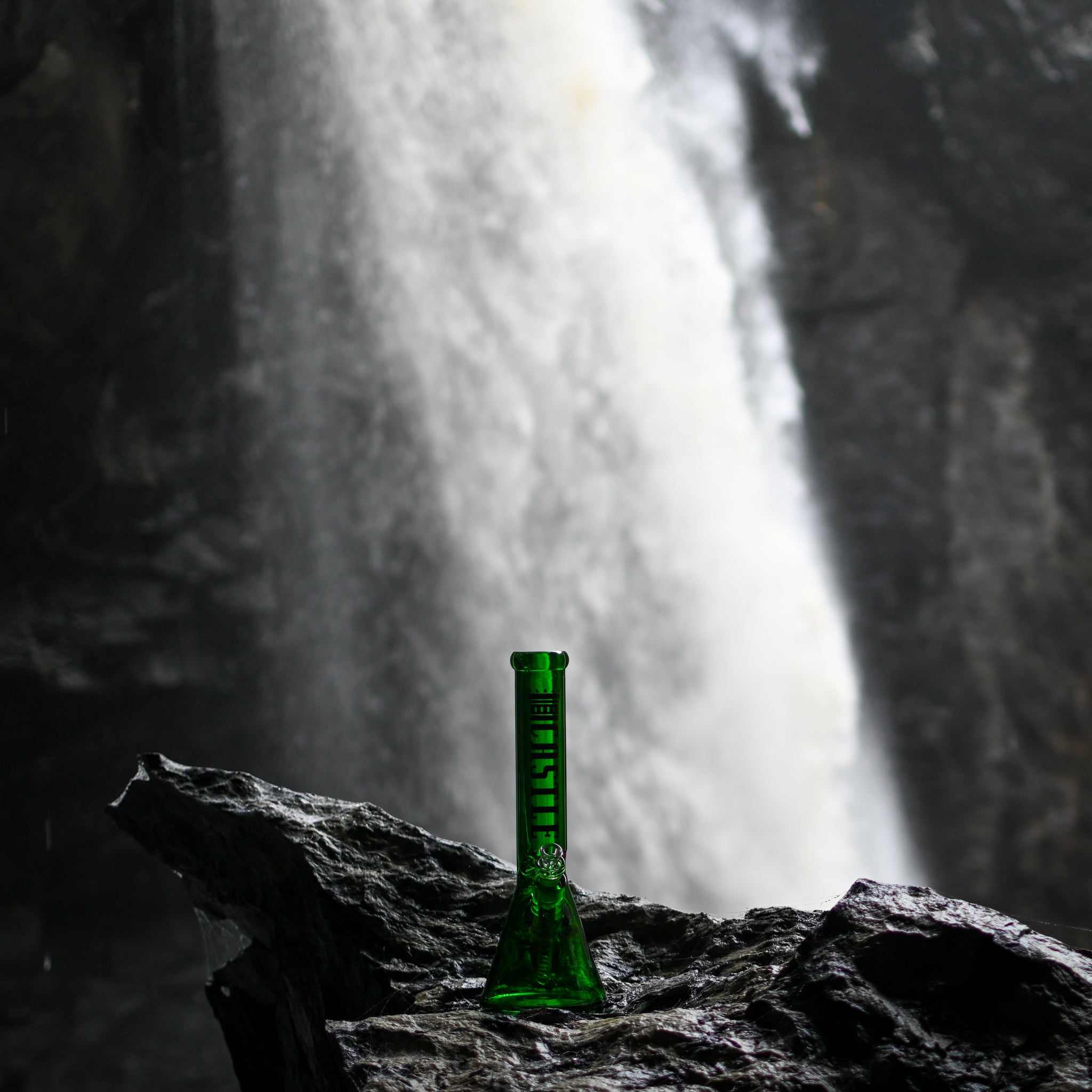 A bright green Castle-branded beaker bong resting on a dark rocky ledge, with a tall waterfall cascading in the blurred background.