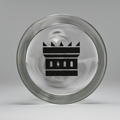 Close-up image of the bottom of a beaker bong, showing the Castle Glassworks logo prominently displayed. The logo features a stylized castle with three windows, etched in black on the clear glass base, set against a soft grey background.