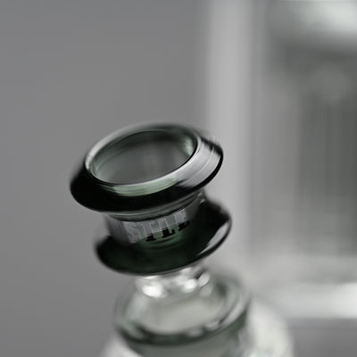Close-up image of the bowl of a beaker bong, highlighting the rim and neck with the Castle Glassworks logo. The bowl is a transparent black, contrasting against the clear glass of the bong and the soft grey background.