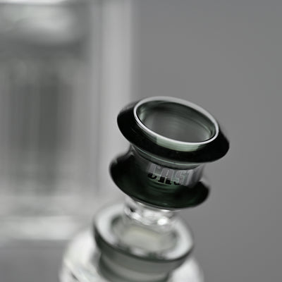 Close-up image of the bowl of a beaker bong, focusing on the rim and neck with the Castle Glassworks logo. The bowl is a transparent black, contrasting with the clear glass of the bong and the soft grey background.