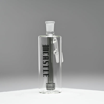 A glass ash catcher with a sleek, minimalist design, featuring the brand name Castle in bold black letters on the side. The ash catcher is equipped with a black matrix percolator. It is set against a smooth, grey background that highlights the clean lines and transparency of the glass. The image emphasizes the modern and sophisticated look of the ash catcher, with the branding and design elements standing out clearly.