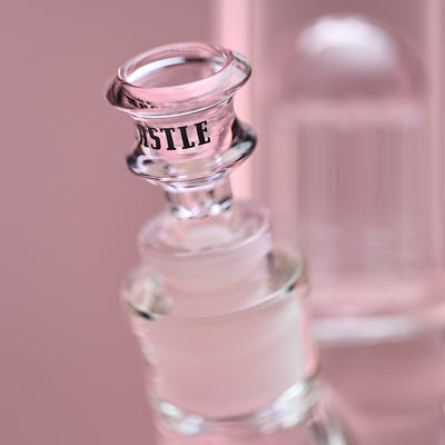 Close-up view of the bowl piece on a clear glass beaker bong with pink accents, set against a soft pink background. The image highlights the "CASTLE" logo on the bowl, showcasing the attention to detail and branding of the glass accessory. The background subtly blurs, focusing the viewer's attention on the bowl and the upper part of the percolator.