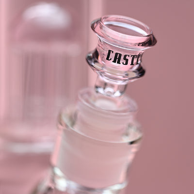 Close-up view of the bowl on a glass beaker bong with pink accents, set against a soft pink background. The image highlights the "CASTLE" logo on the bowl, emphasizing the branding and craftsmanship of the glass accessory. The background subtly blurs, focusing the viewer's attention on the bowl and part of the percolator in the distance.