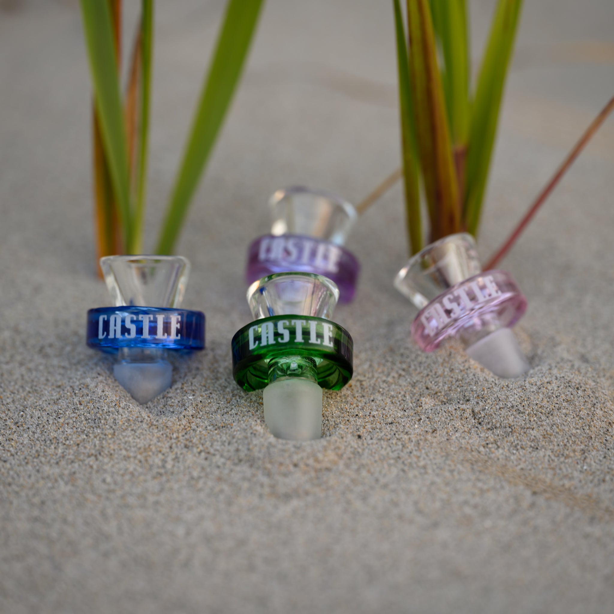 A set of four vibrant glass bong bowls from Castle Glassworks is arranged in the soft sand, partially nestled among blades of beach grass. Each bowl features a bold, colored rim with the "CASTLE" branding in white, showcasing a mix of deep blue, rich green, and soft purple hues. The crystal-clear glass and frosted joints highlight their high-quality craftsmanship. The natural setting, with warm sand and fresh greenery, contrasts beautifully with the smooth, polished glass, emphasizing aesthetic appeal.