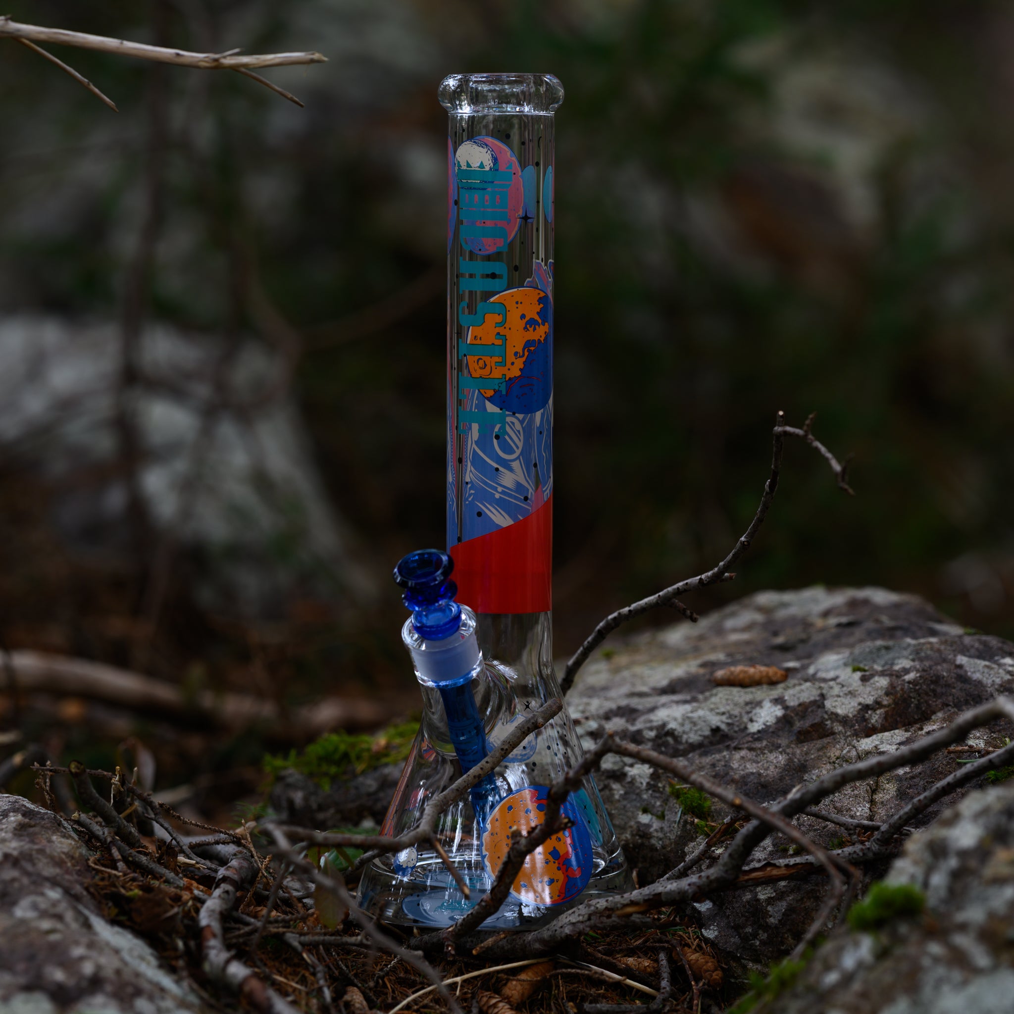 Clear glass water pipe with colorful space-themed artwork on its neck, resting on rocks among branches in an outdoor setting.